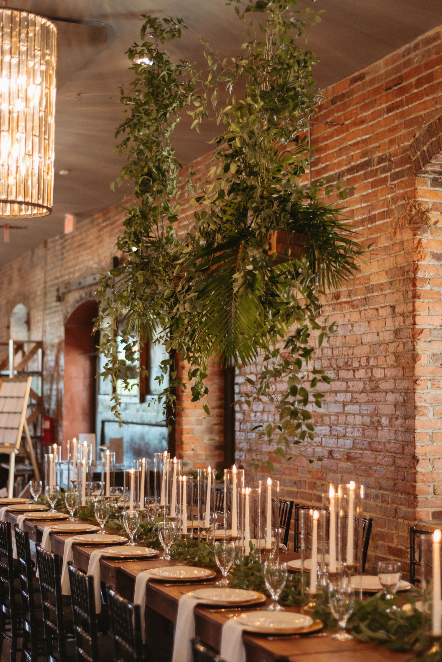 Elegant wedding reception table setting with greenery and candles against exposed brick walls.