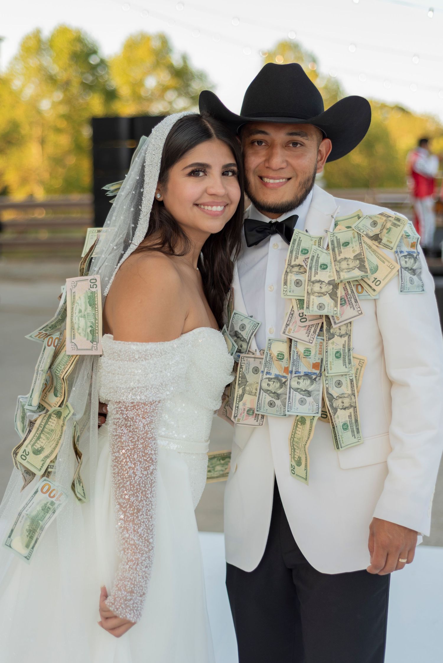 A couple in western wedding attire pose with money pinned to their clothing during an outdoor celebration.