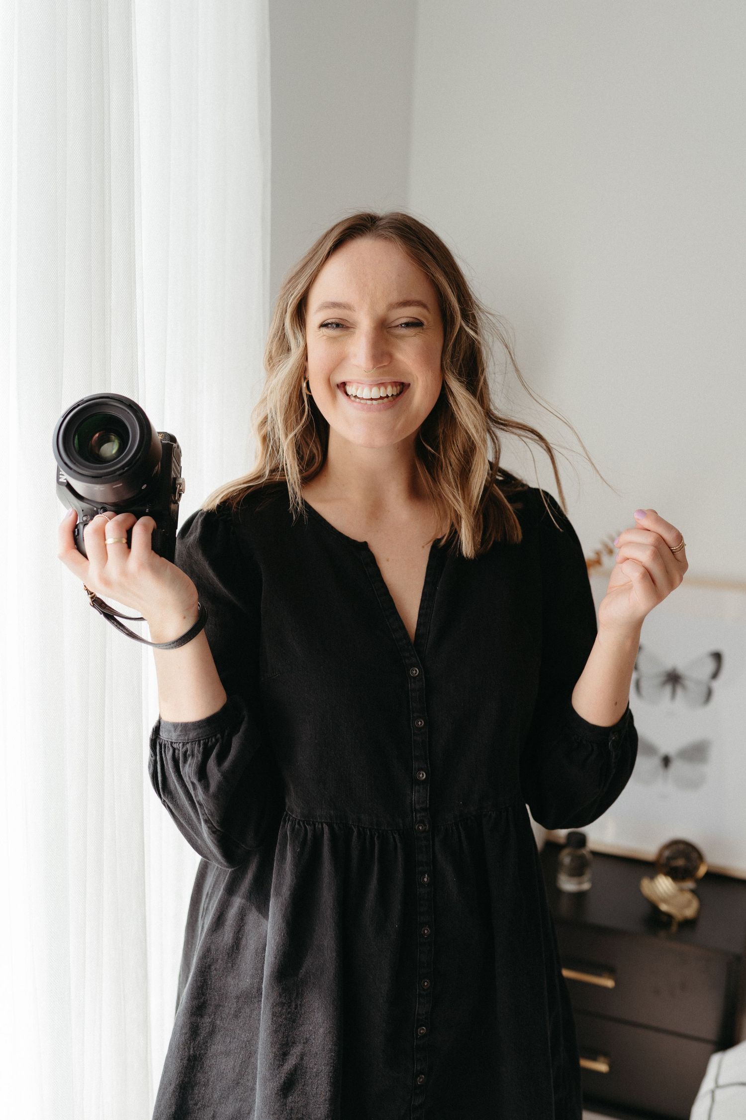 Photographer Rhesa Rogers in a black button-up shirt smiles while holding a camera in a bright, minimalist interior setting.