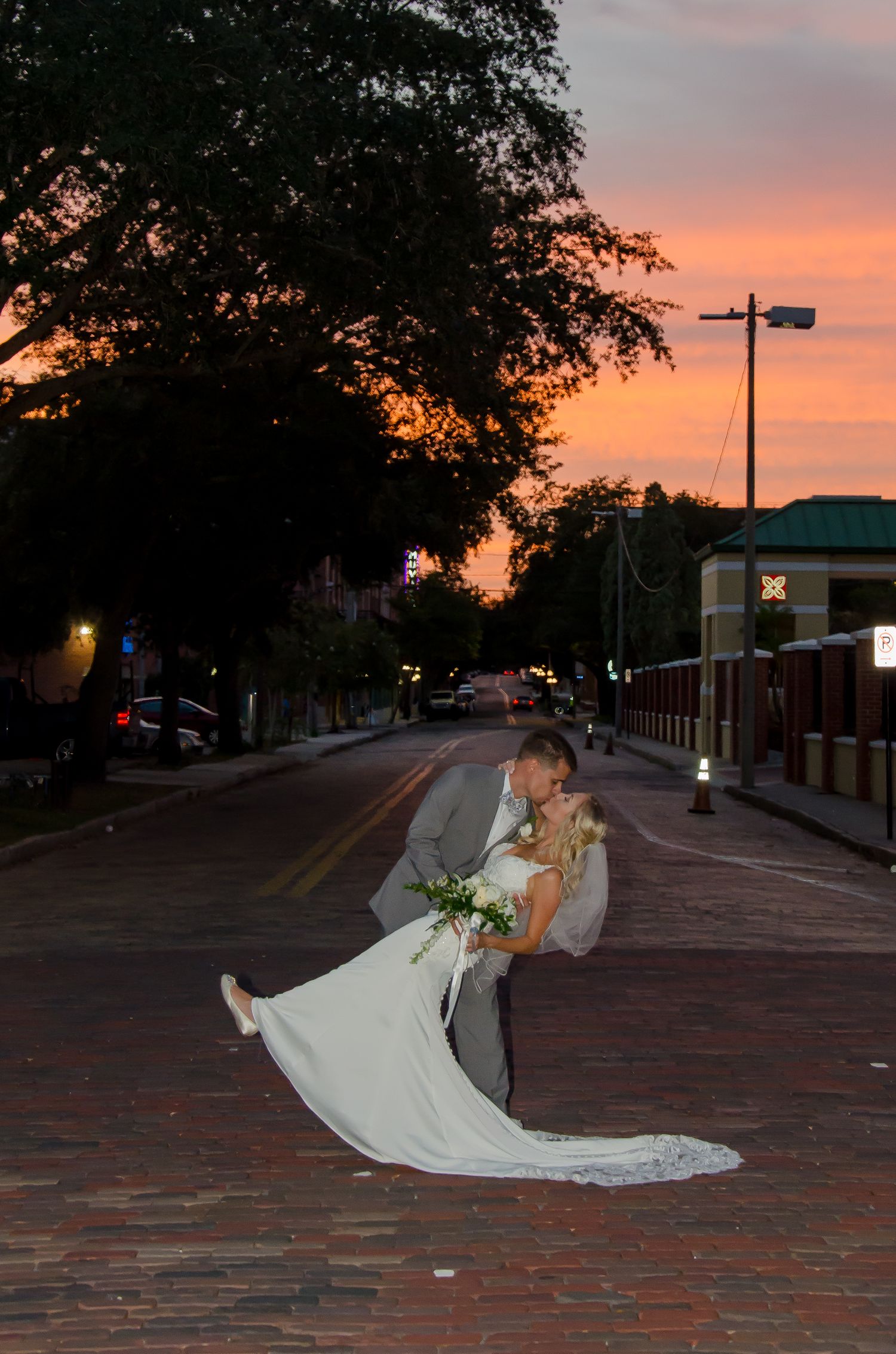 Bride and groom share a romantic dip kiss on a brick street at sunset with a vibrant orange sky backdrop.