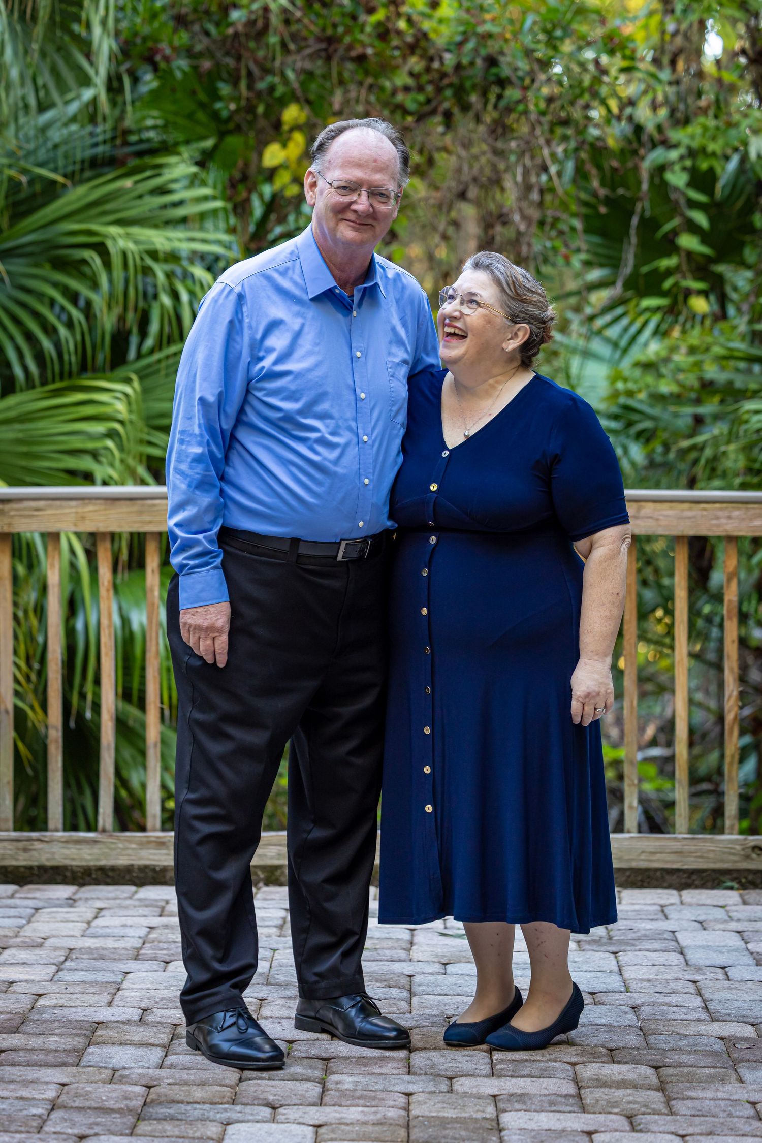 Smiling older couple posing together on a brick patio surrounded by lush tropical greenery.