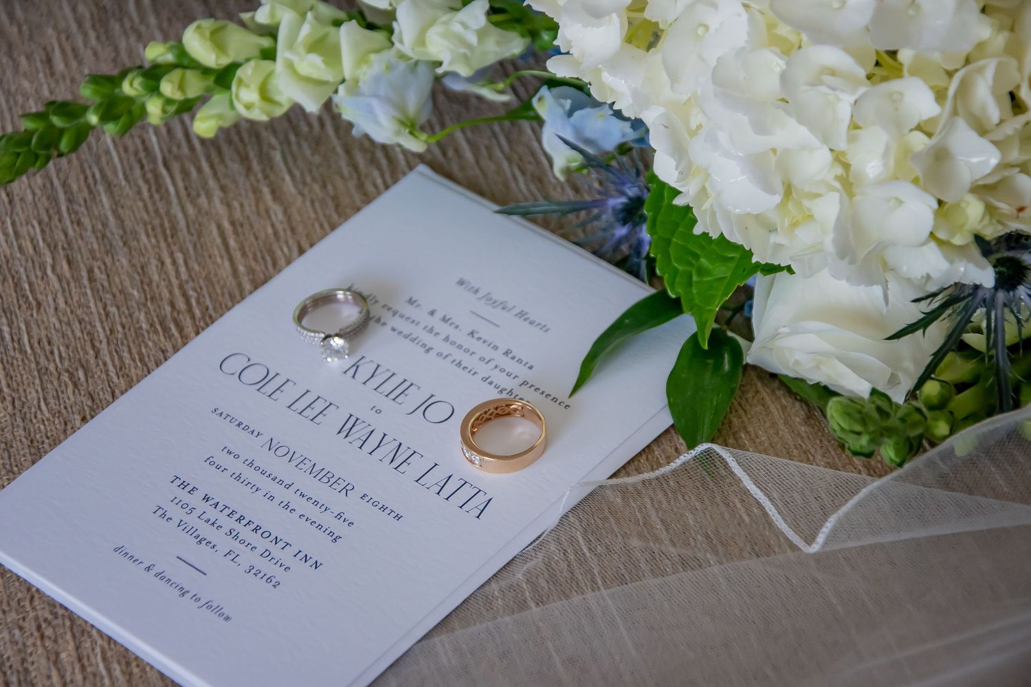 Wedding invitation with gold rings and diamond ring resting on white hydrangea bouquet on wooden surface.