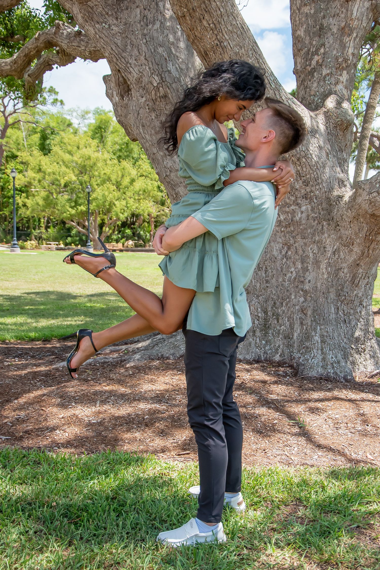 A man lifts a woman in a romantic embrace near a large tree in a sunny park, both wearing matching sage green outfits.