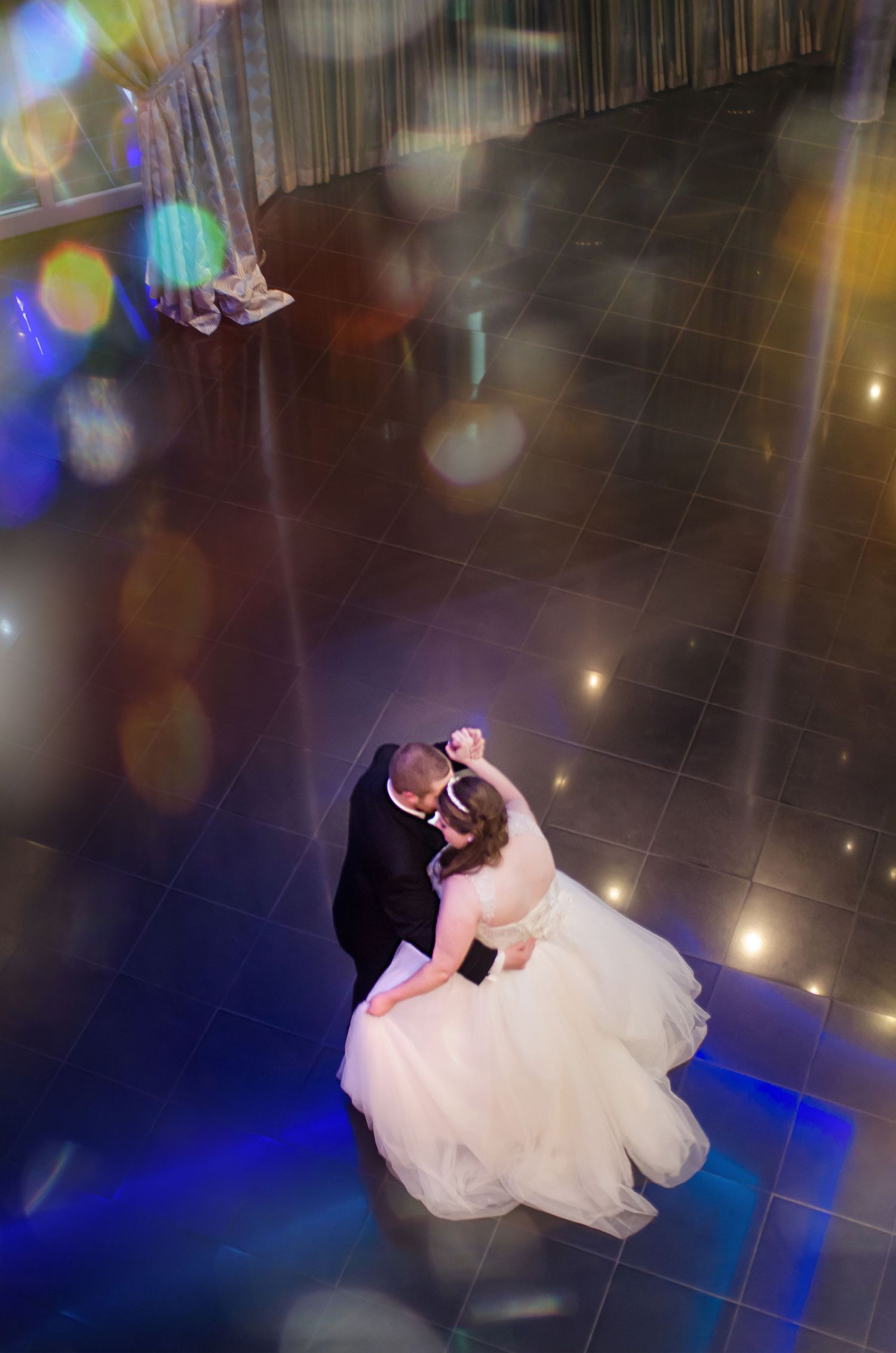 Aerial view of bride and groom sharing their first dance on a dark tile floor with colorful bokeh lights at wedding reception.