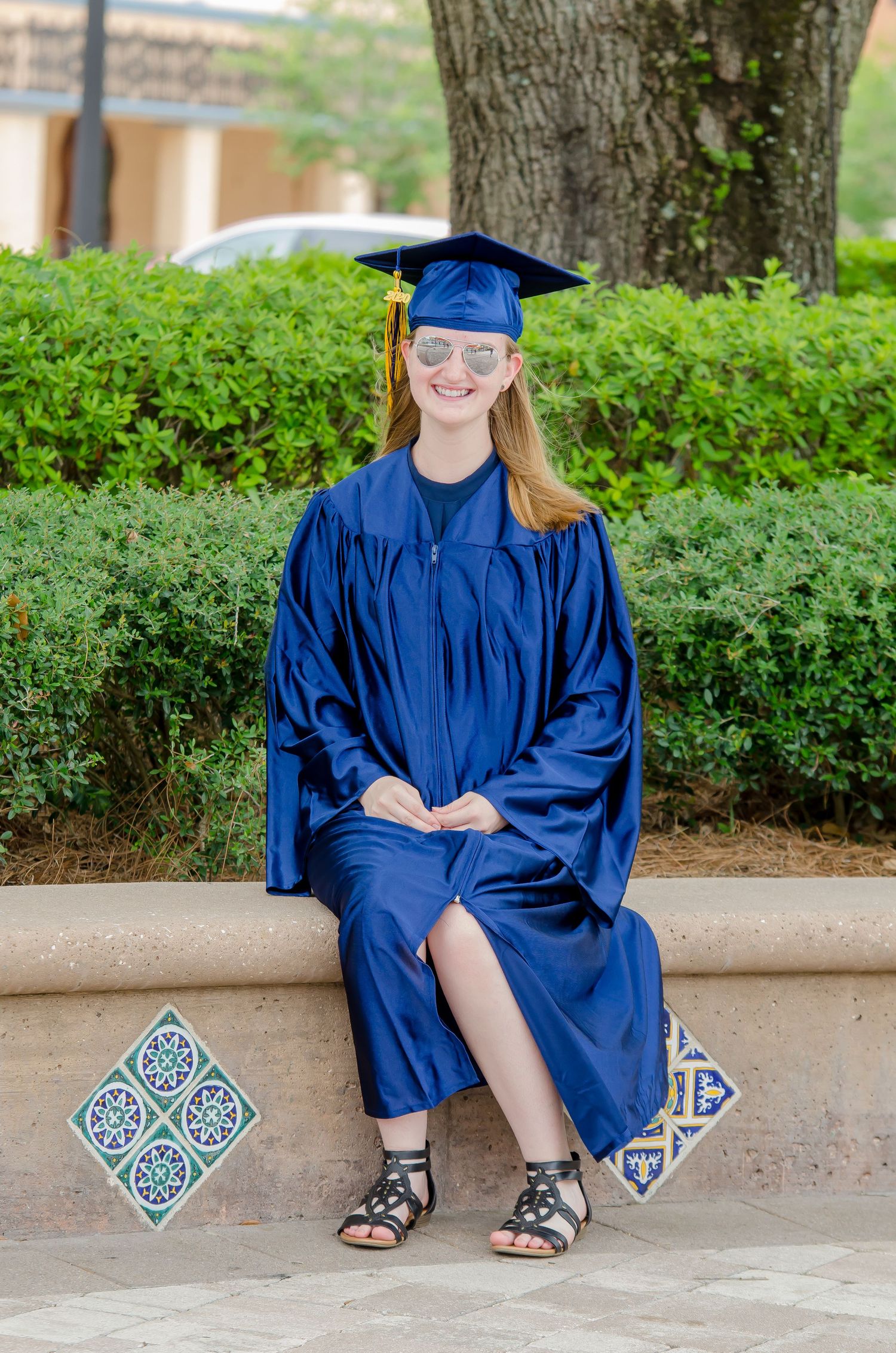Young woman in blue graduation cap and gown sitting outdoors, smiling with sunglasses on a decorative tiled bench.