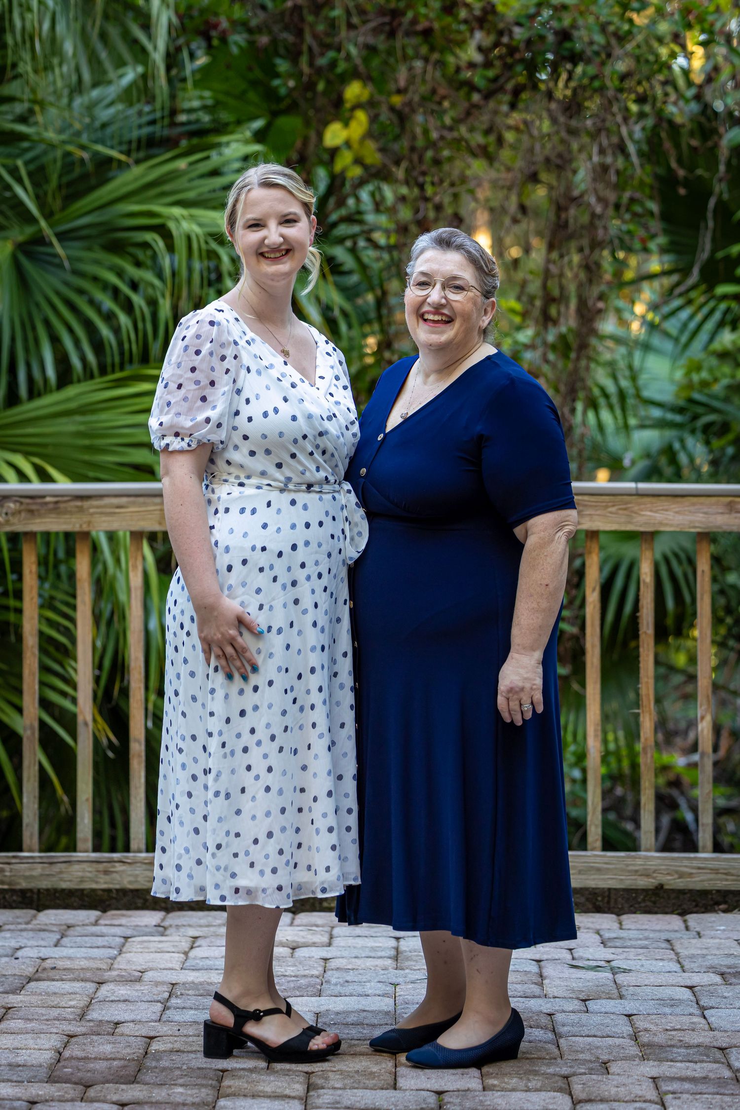 Two smiling women in dresses pose together on a patio surrounded by lush tropical greenery.