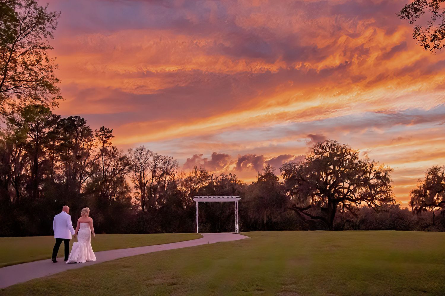 Bride and groom walk hand-in-hand toward a garden arch beneath a stunning fiery sunset sky.
