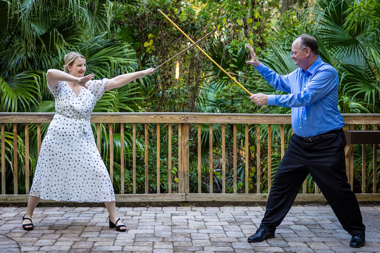 A man and woman playfully duel with bamboo sticks on a garden patio surrounded by lush tropical greenery.