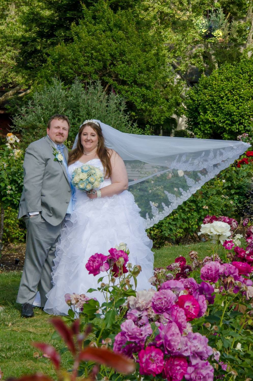 Bride and groom pose among vibrant pink roses in a garden, her veil flowing in the breeze on their wedding day.