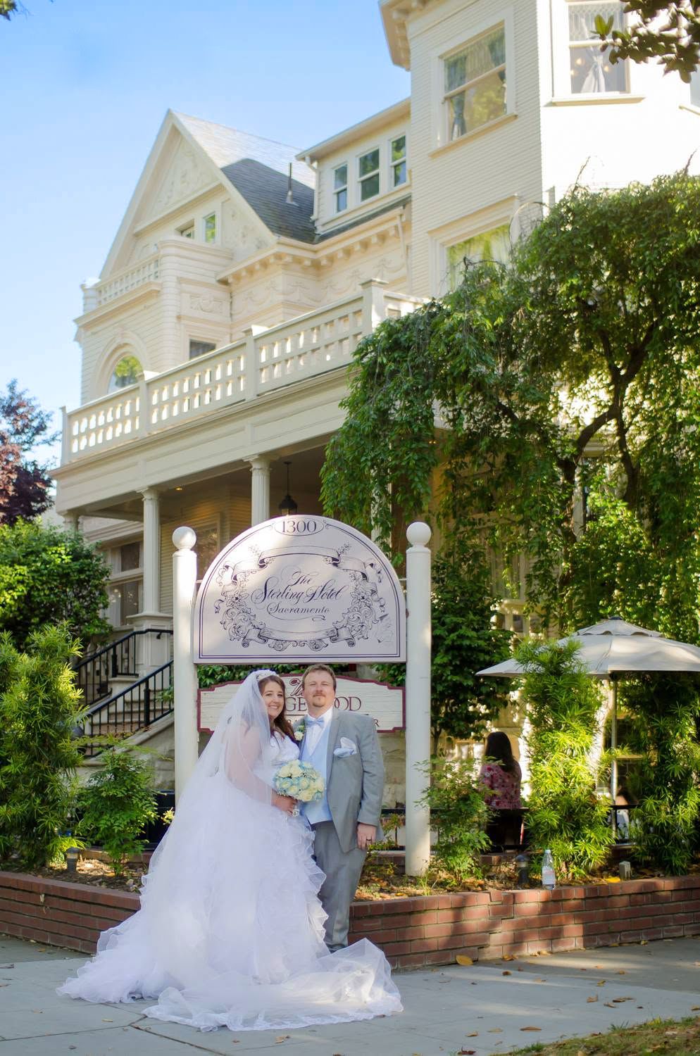 Bride and groom posing in front of elegant Victorian wedding venue with lush gardens on a sunny day.