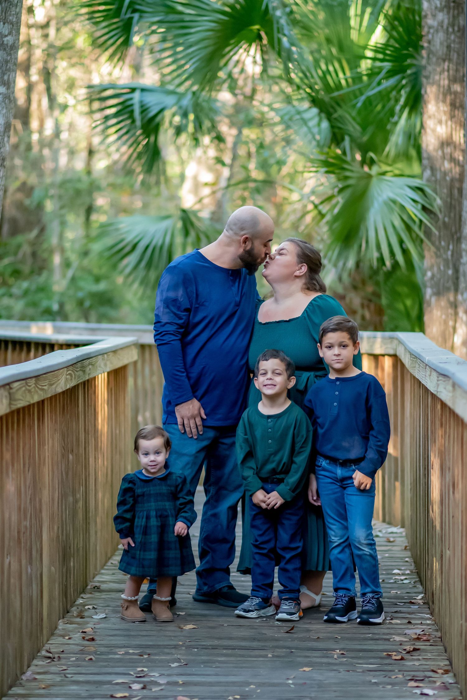 Family of five poses on a wooden boardwalk surrounded by lush greenery, parents kissing while three children stand in front.