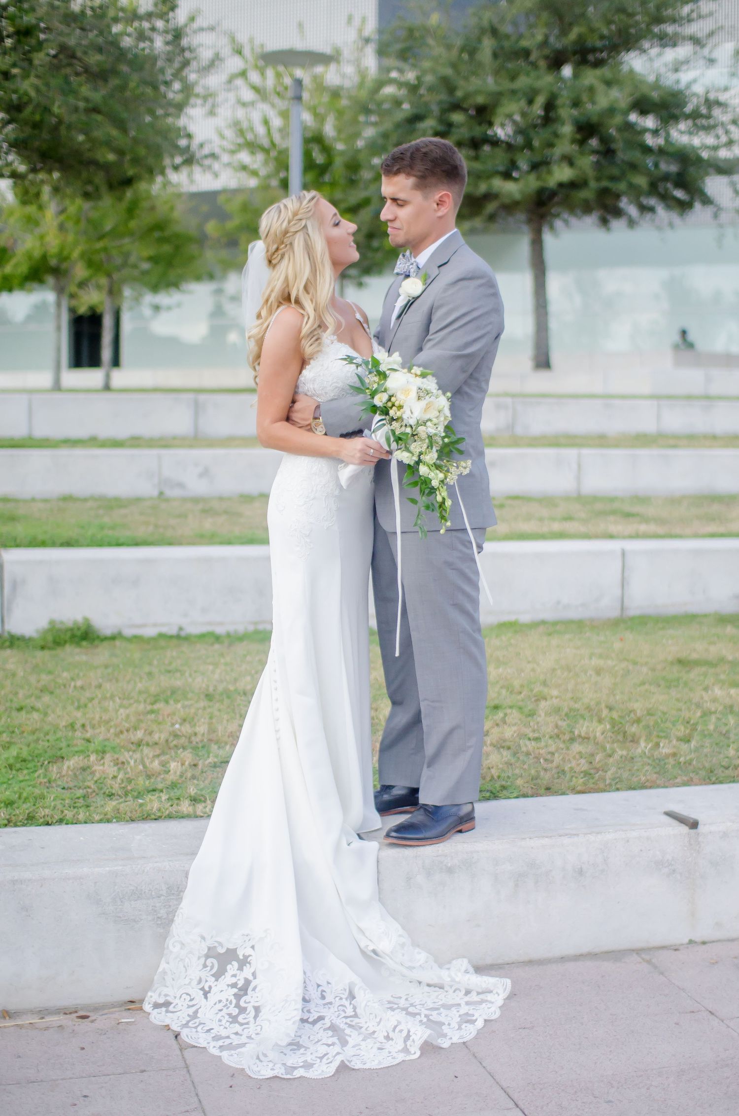 Bride in white lace gown and groom in gray suit share romantic moment outdoors holding white floral bouquet.