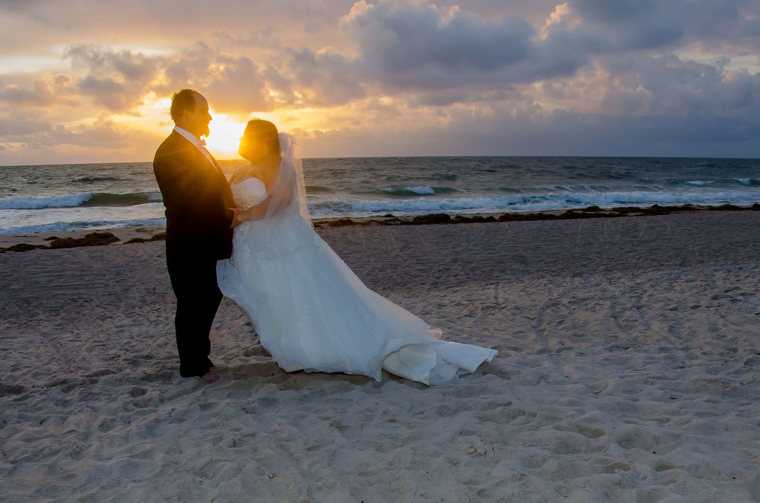 Newlywed couple embracing on a sandy beach at sunset with ocean waves in the background.