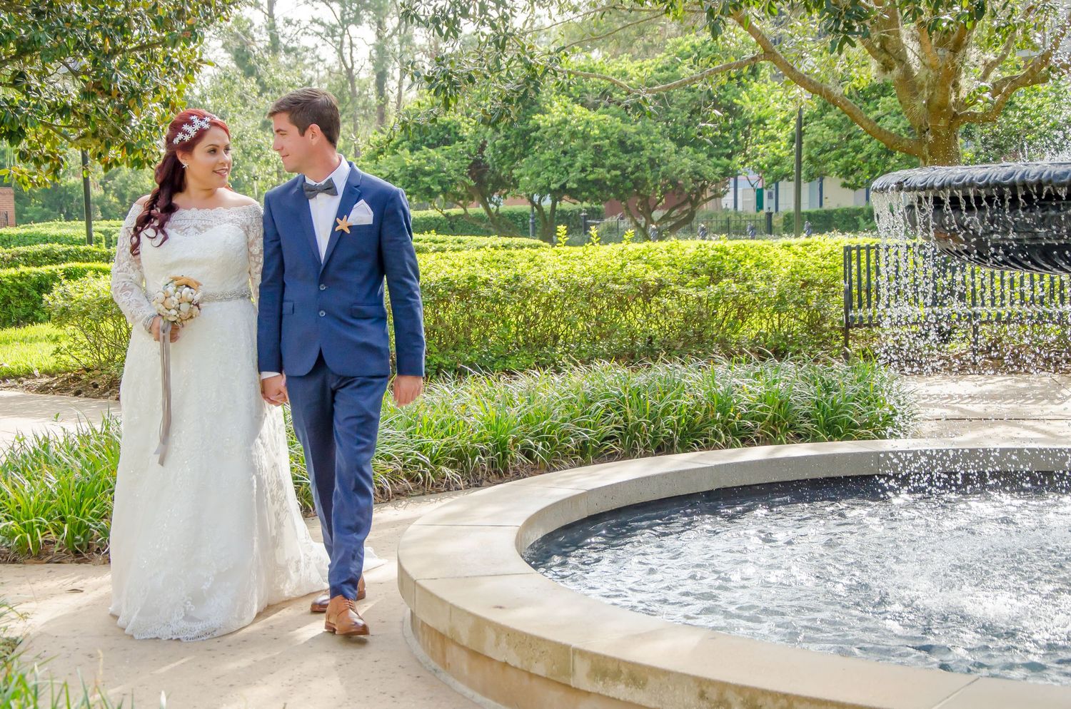 Bride and groom walking hand in hand beside a fountain in a lush garden on their wedding day.