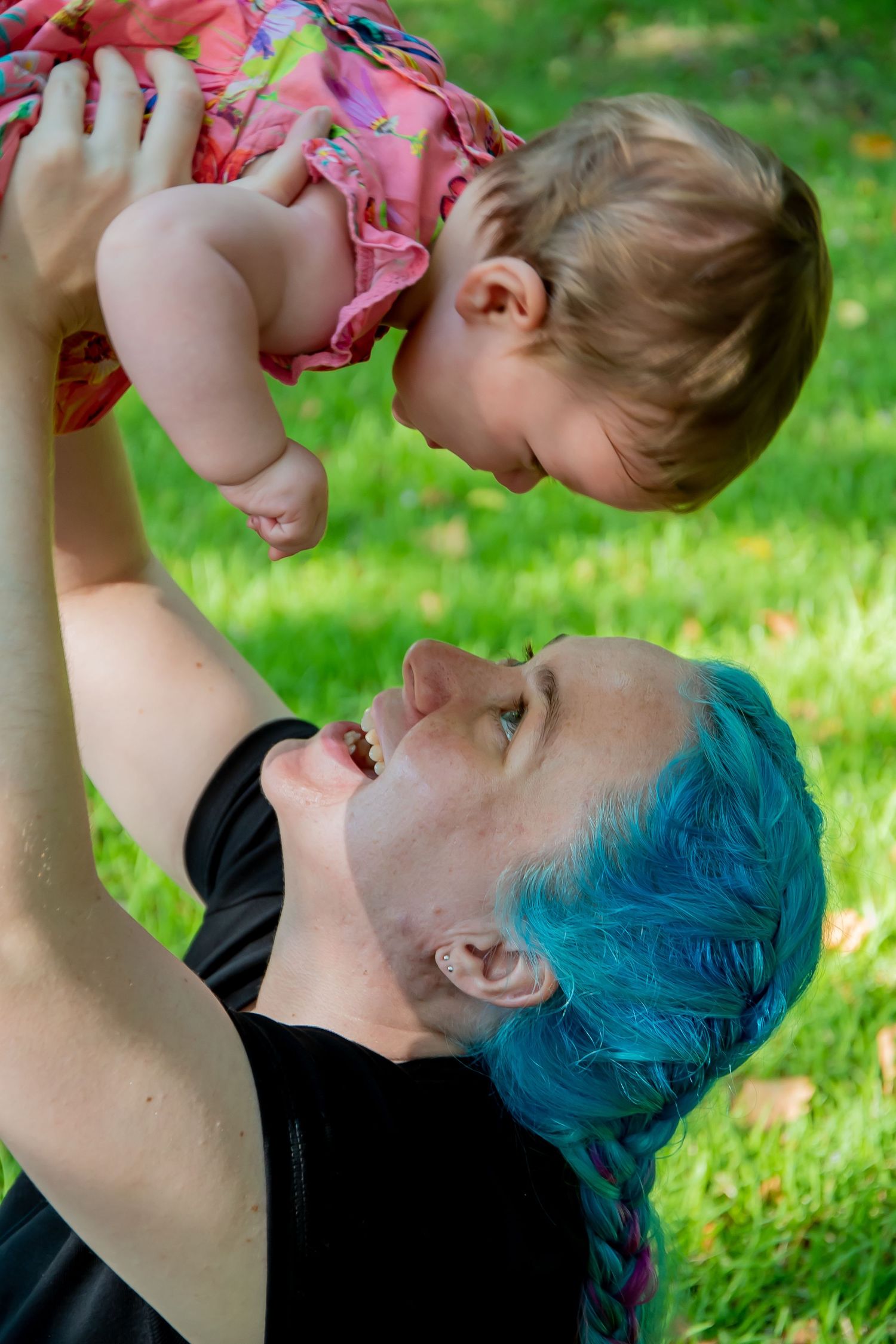 A woman with teal braided hair lifts a smiling baby in a pink dress outdoors on green grass.