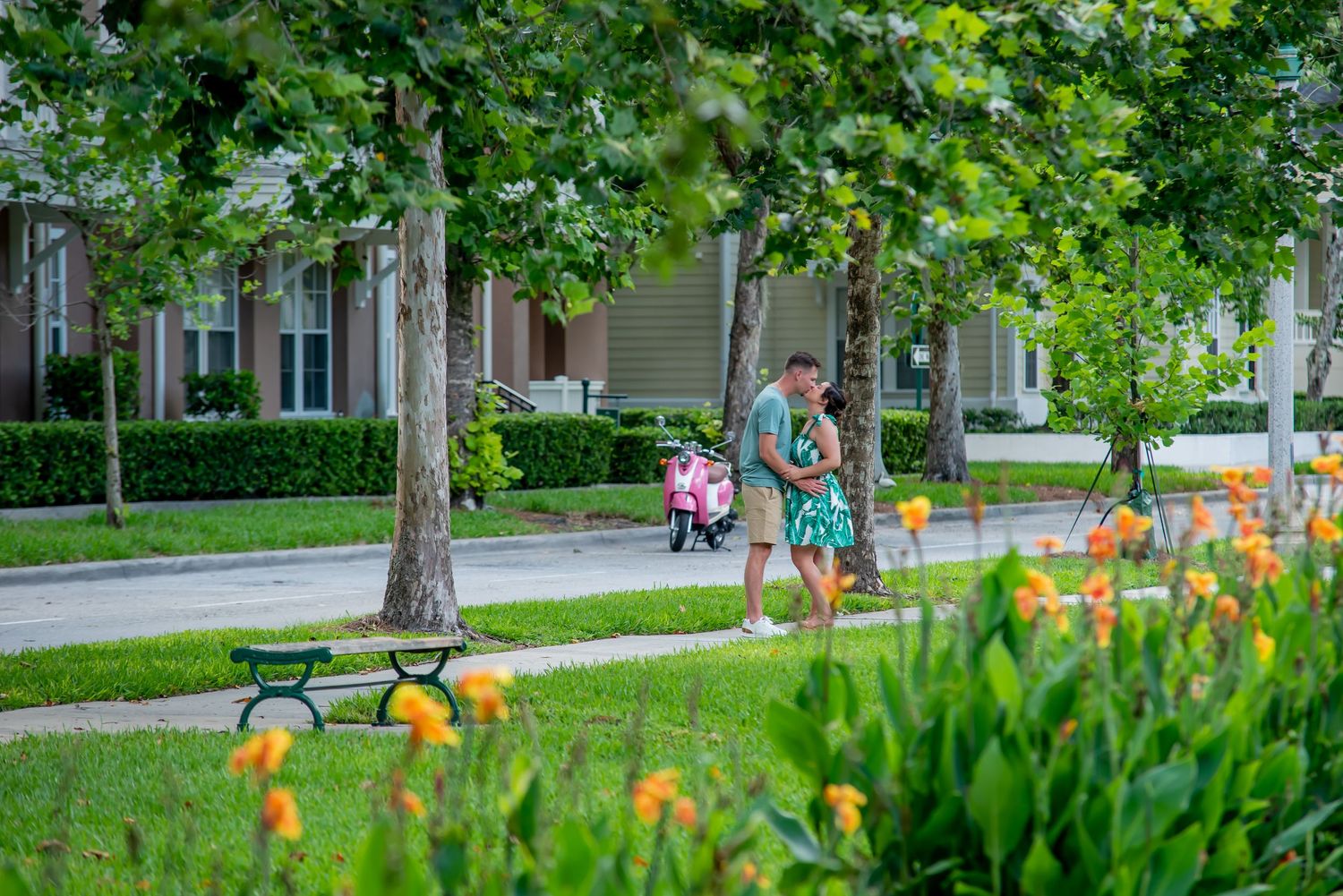 Family strolling on a tree-lined sidewalk past colorful flowers and green lawns in a suburban neighborhood.