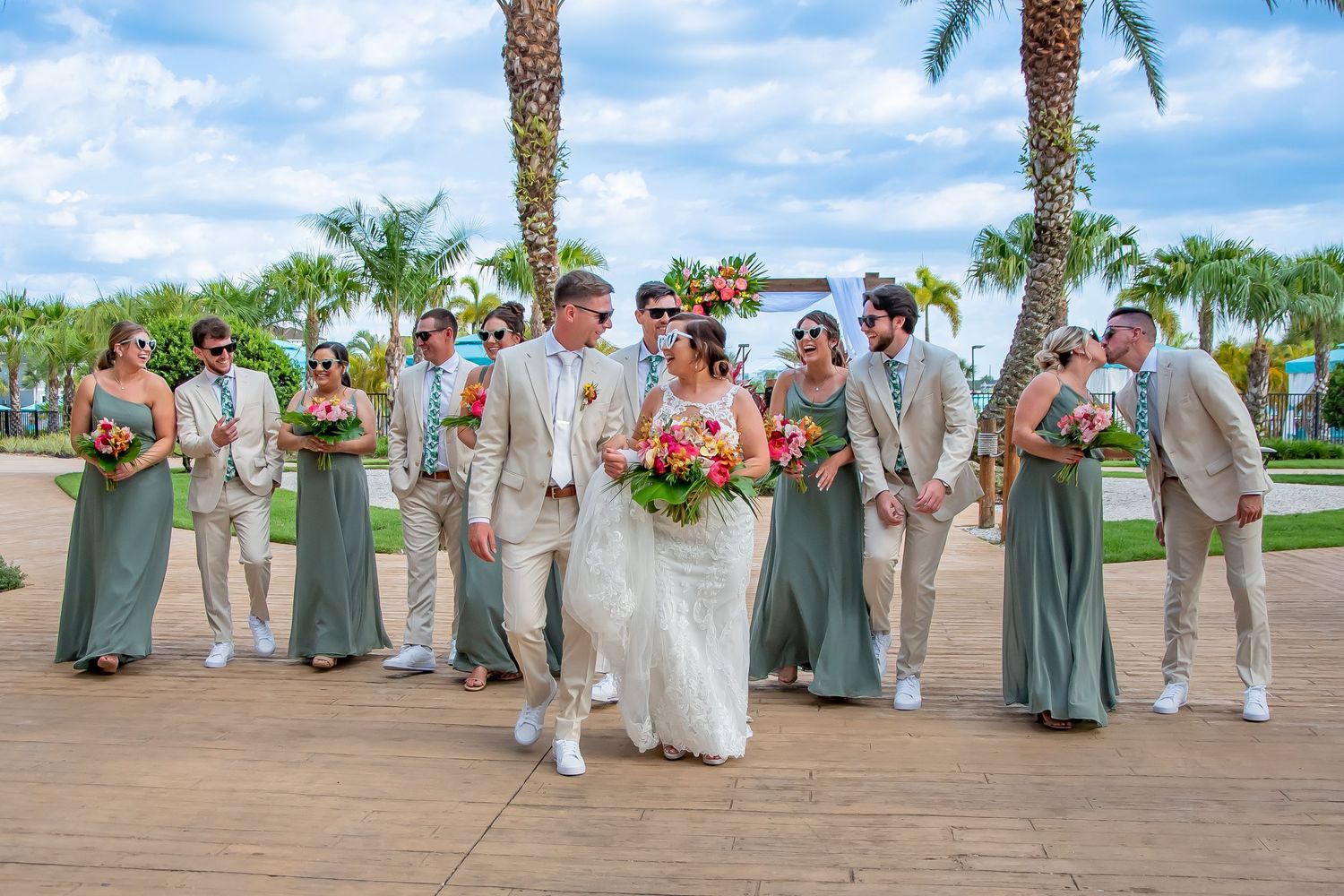 Wedding party walks outdoors near palm trees, bridesmaids in sage green gowns, groomsmen in tan suits, bride and groom centered.