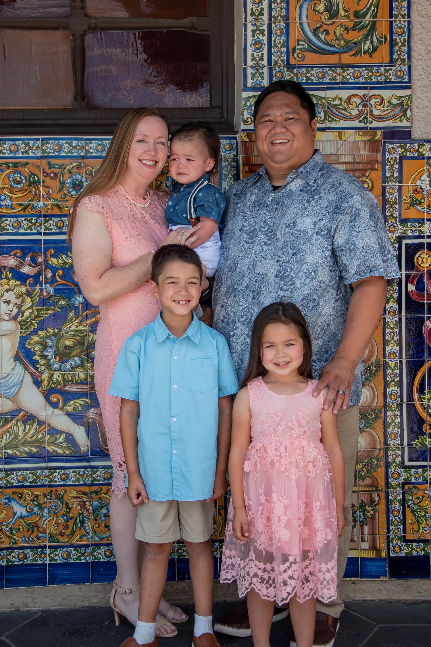 Family of five posing in front of colorful decorative tiled wall, parents with three young children dressed in pastel outfits.