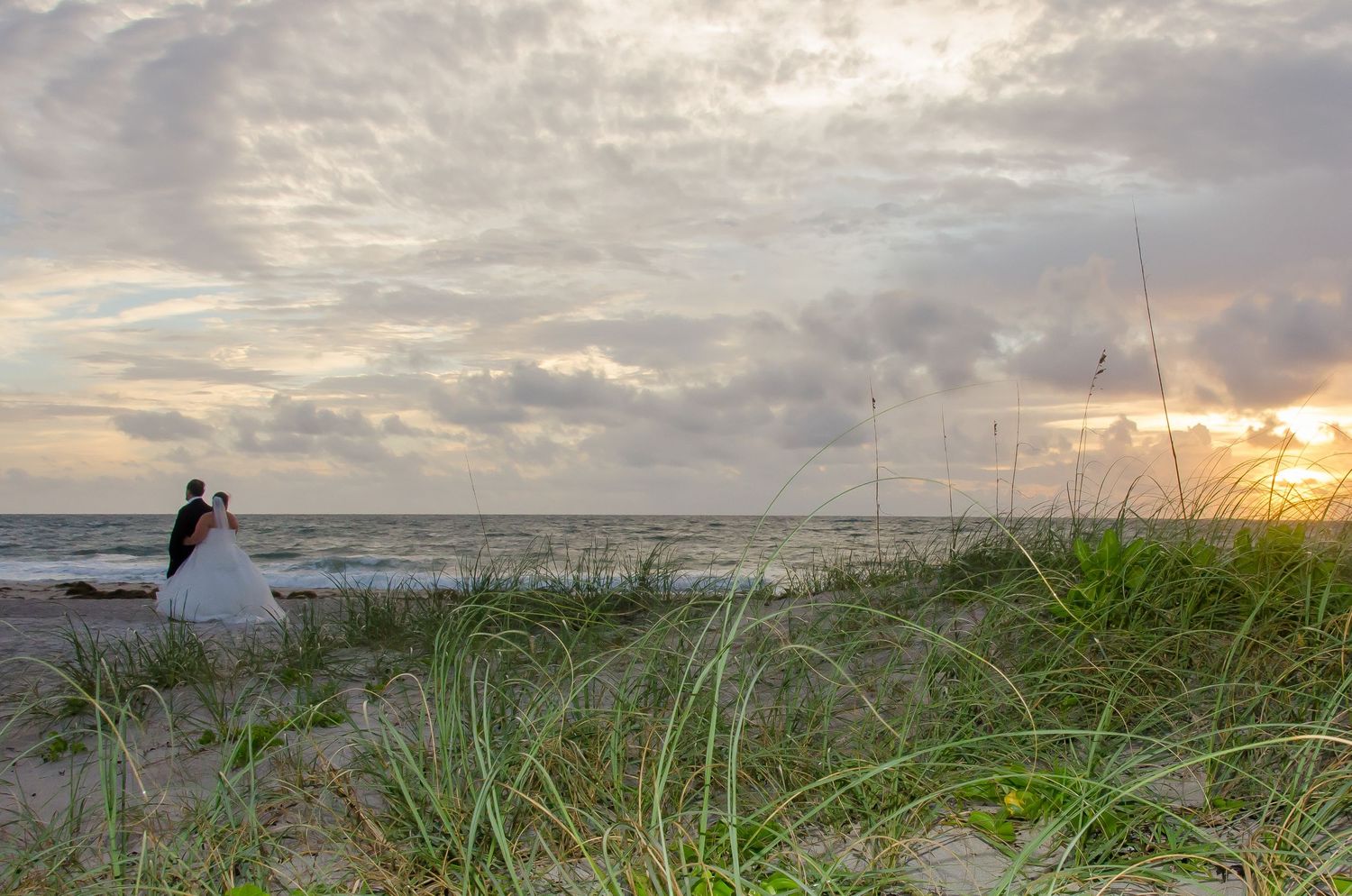 Couple in wedding attire embracing on a sandy beach at sunset with sea grass and dramatic cloudy sky.