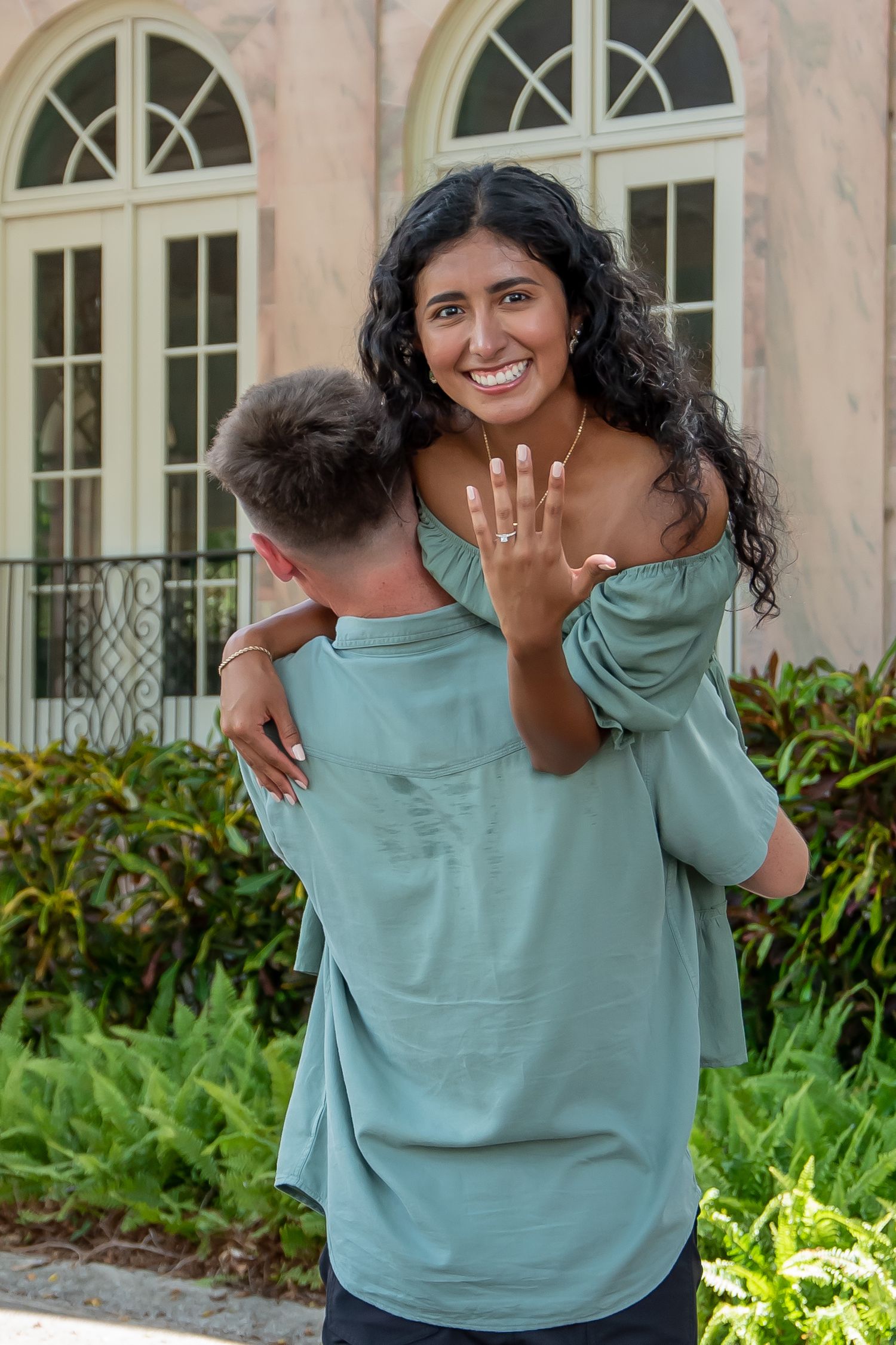 Happy woman shows off engagement ring while embracing her partner outdoors near a building with lush greenery.