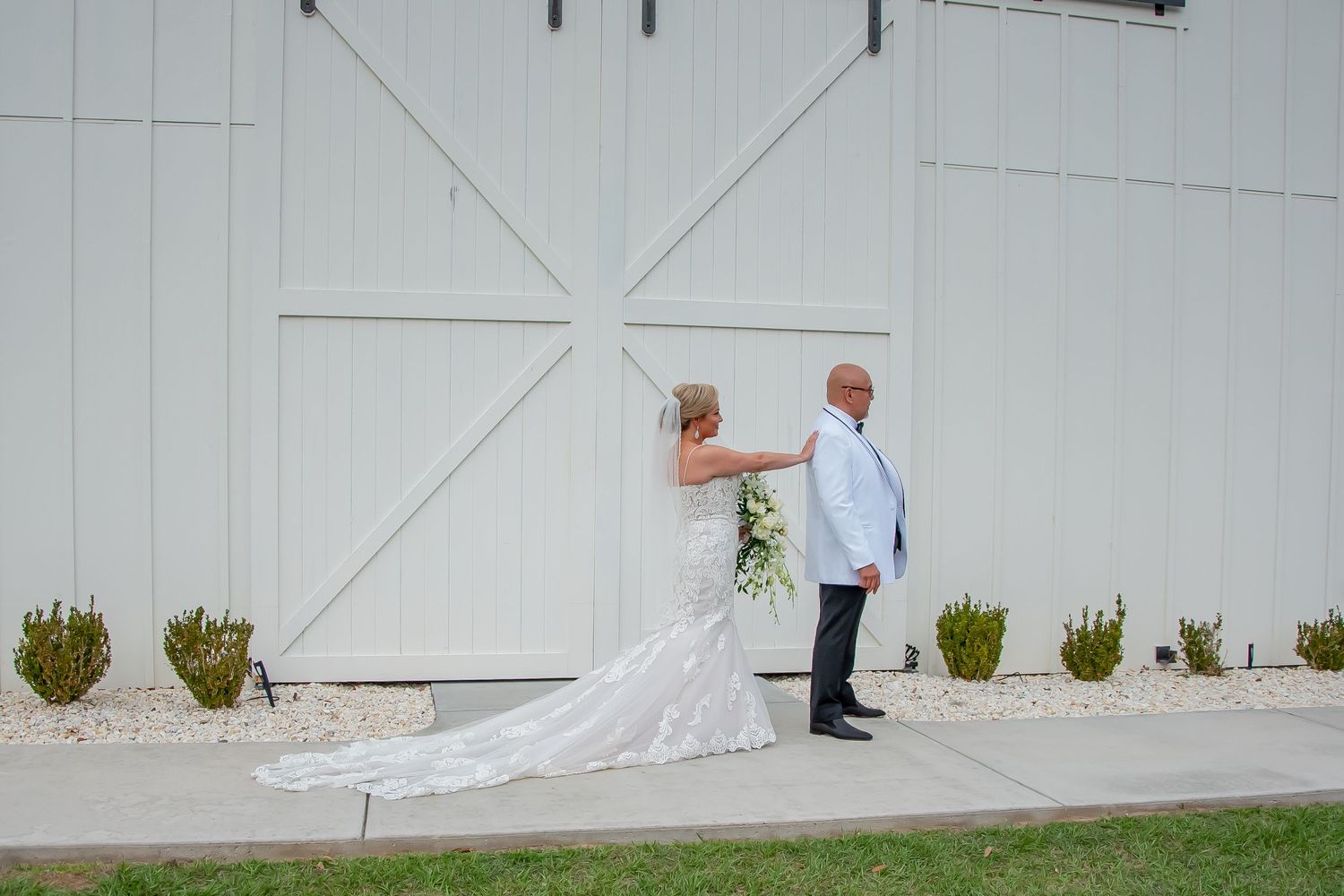 Bride in white gown taps groom's shoulder for first look outside a white barn wedding venue.