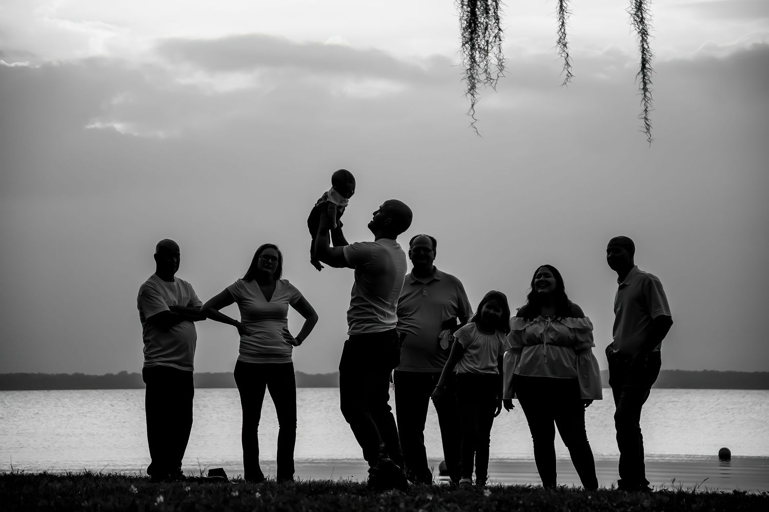 Black and white silhouette of a large family group by a lake, with one person lifting a baby into the air.