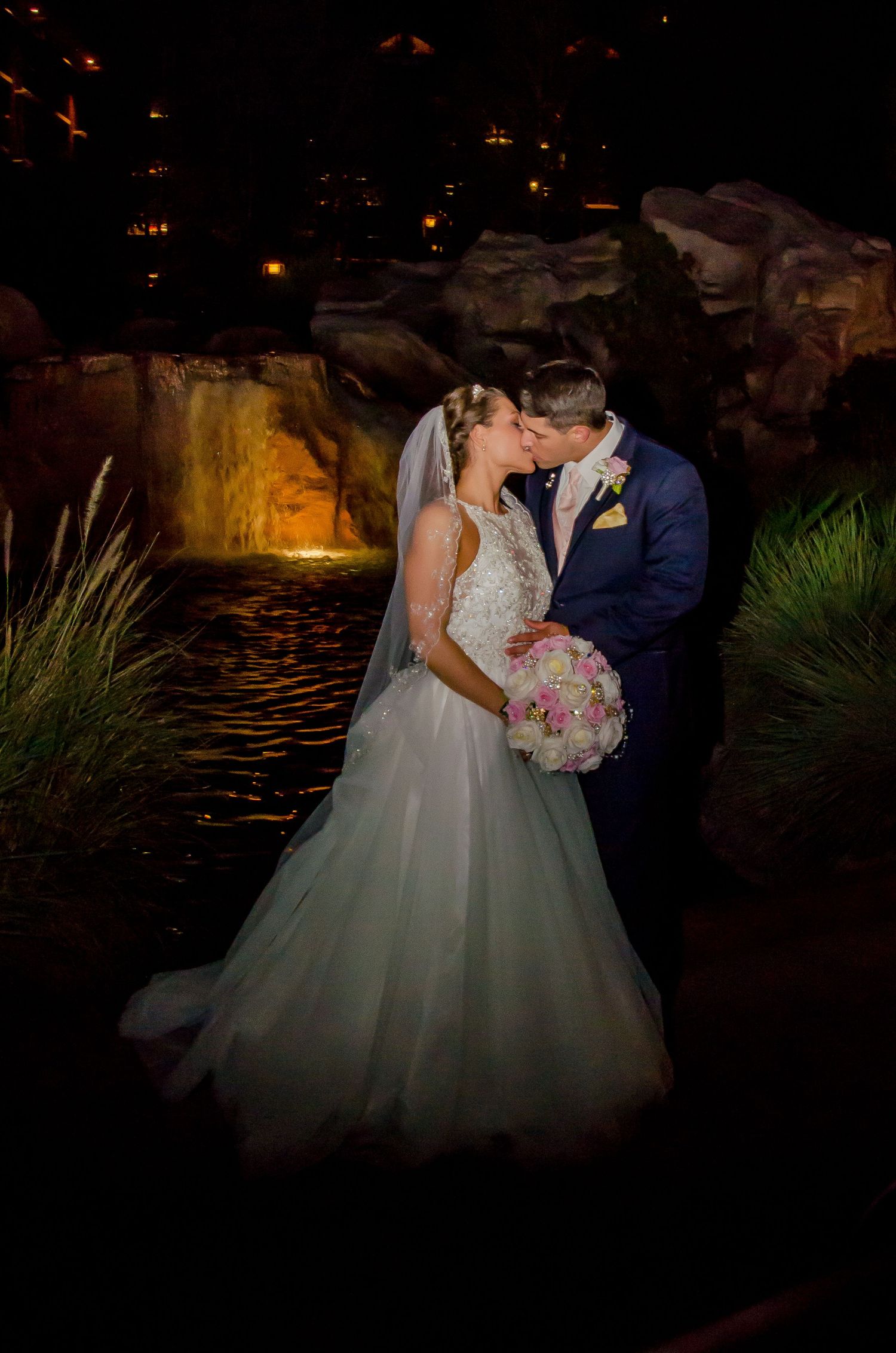Bride and groom share a romantic kiss at night by a waterfall, bride in white gown holding pink bouquet.
