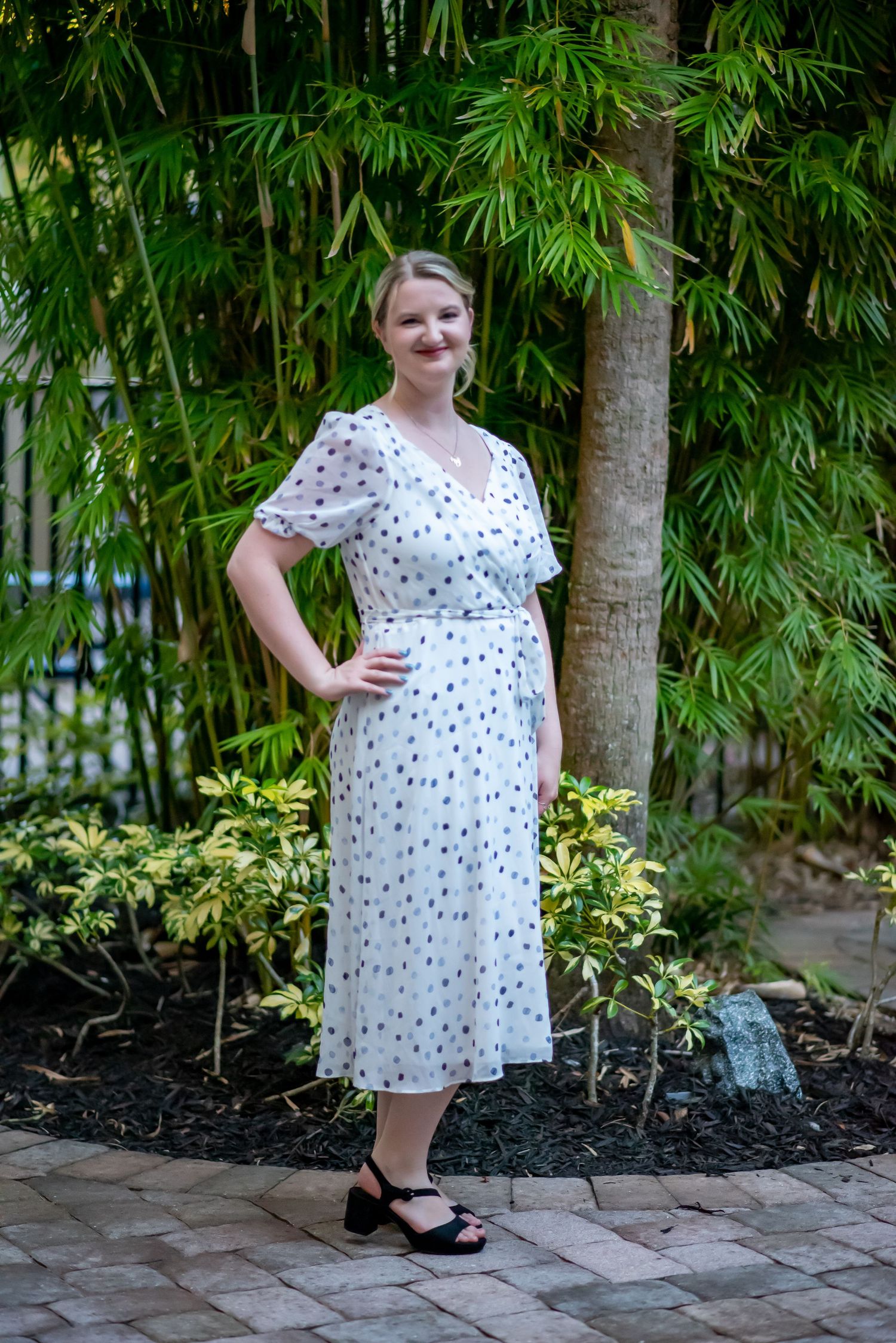 Woman in white polka dot midi dress with flutter sleeves poses in front of lush bamboo garden on brick pathway.