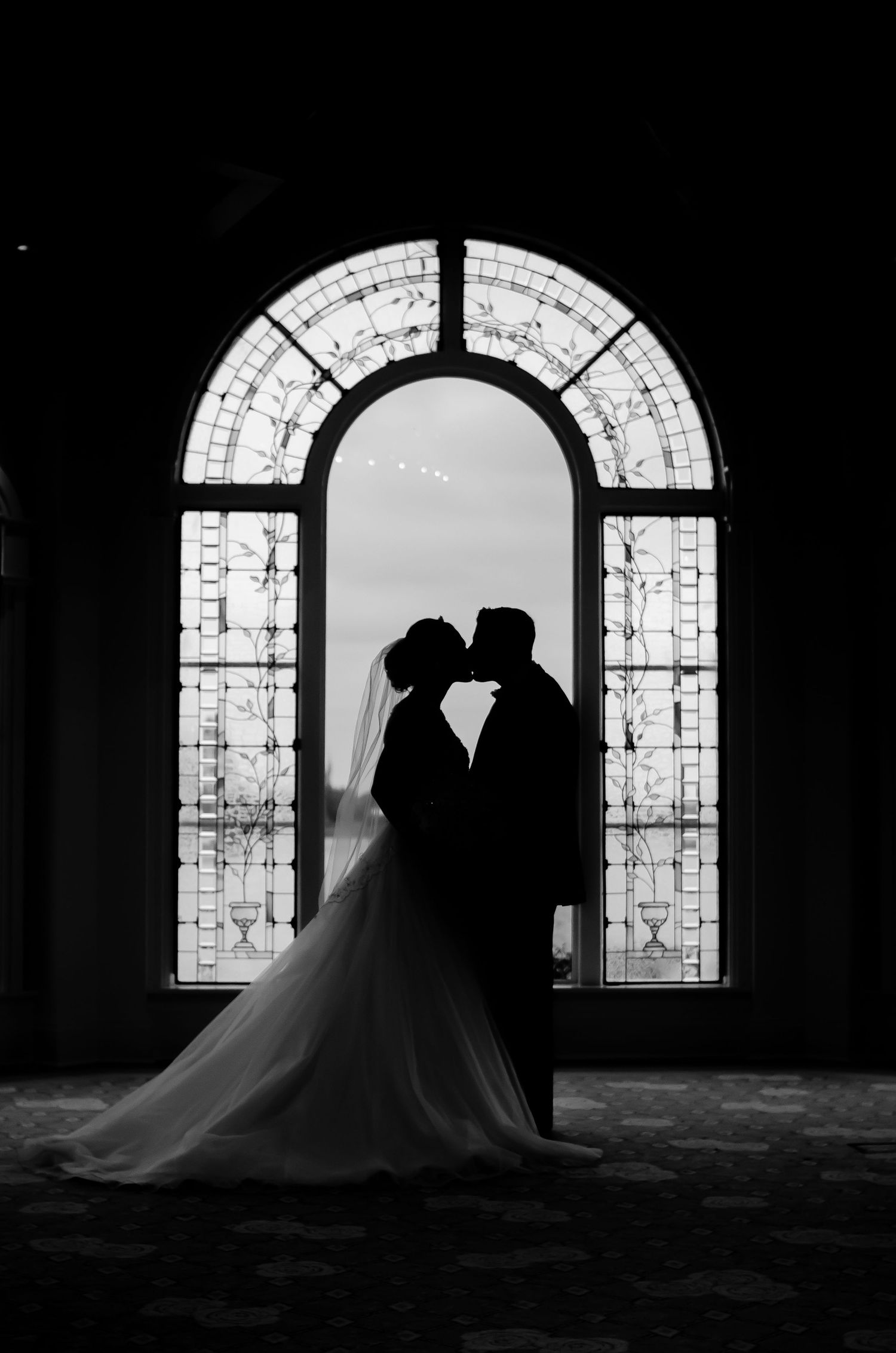 Silhouette of bride and groom kissing in front of ornate arched stained glass window in black and white.