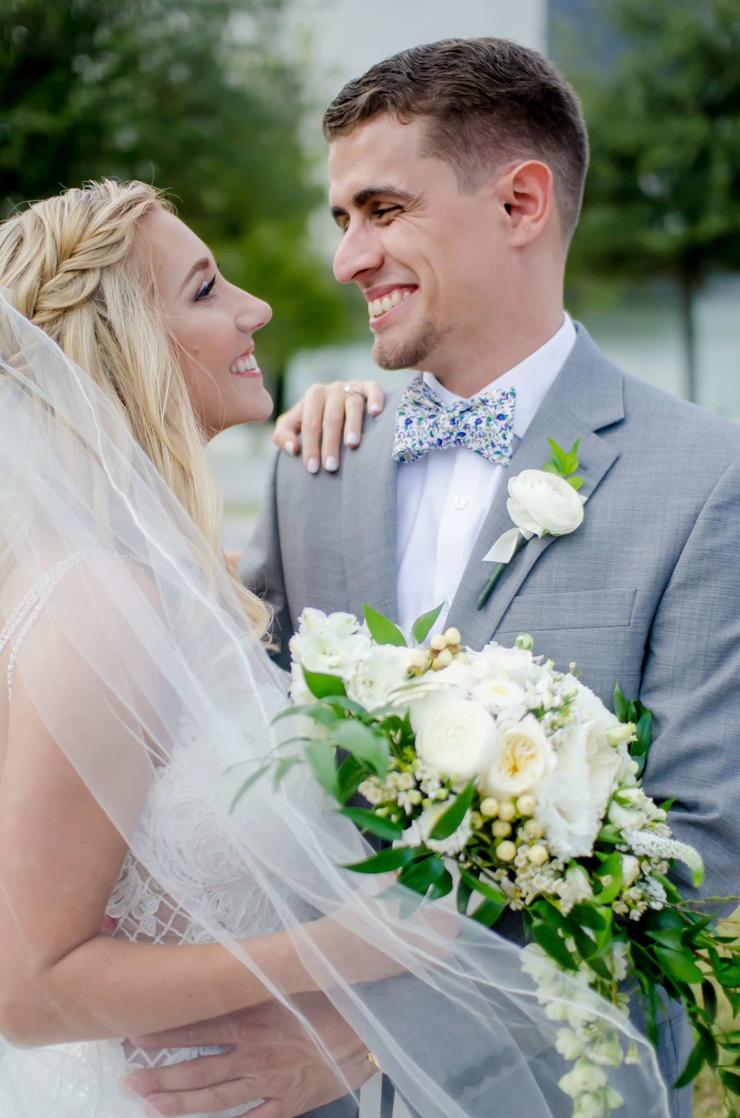 Smiling bride and groom in gray suit with floral bow tie share a tender moment, bride holding white bouquet with greenery.