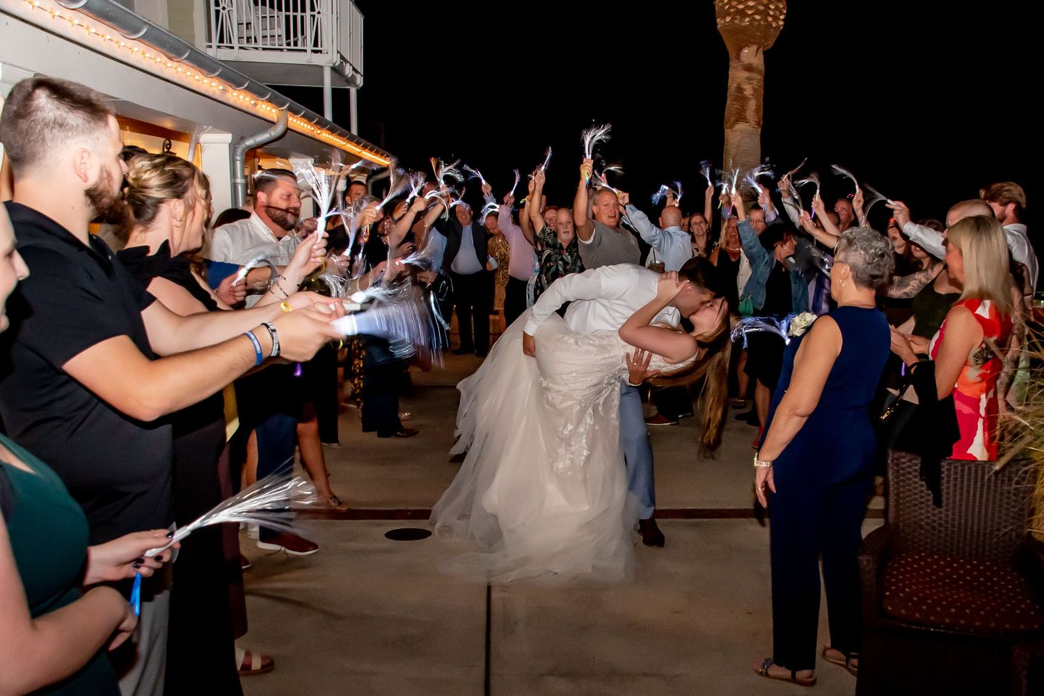 Bride and groom share romantic dip kiss during outdoor wedding sendoff with guests waving sparklers at night.