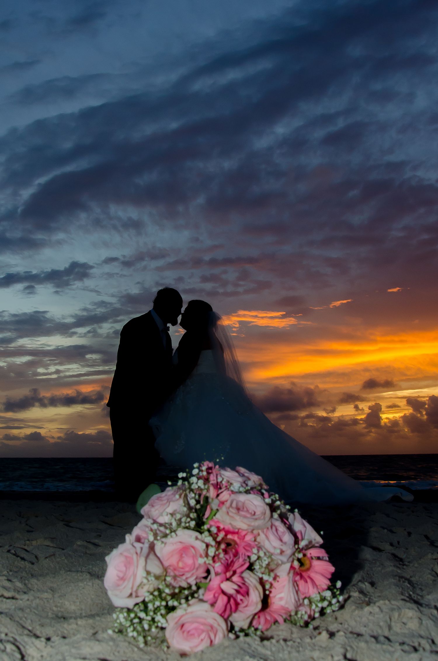 Silhouette of bride and groom kissing at beach sunset with pink rose bouquet in foreground.