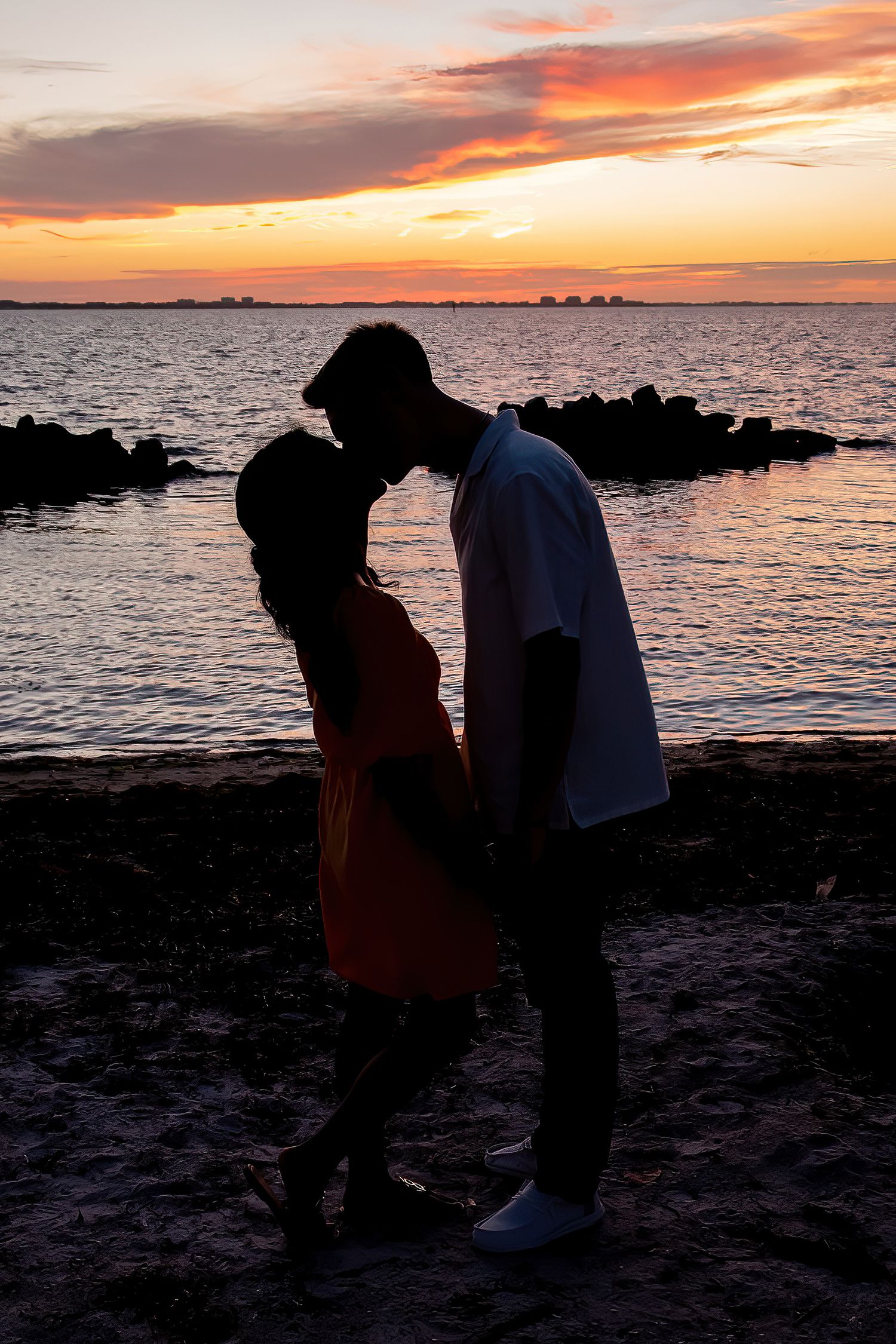 Romantic couple silhouette sharing a kiss on a rocky beach at sunset with vibrant orange and pink sky.
