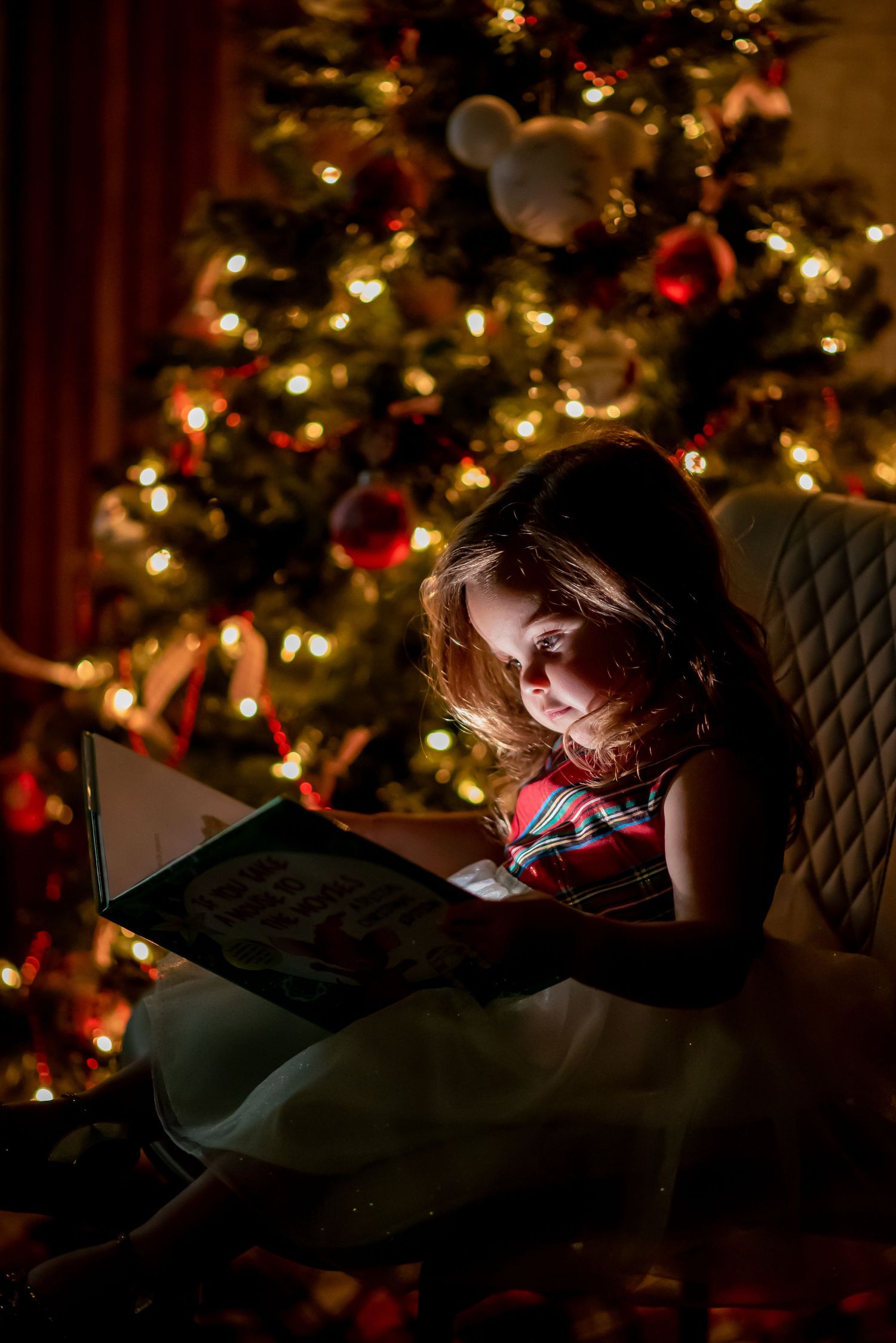 A young girl reads a book by the glow of a decorated Christmas tree with warm golden lights in a dark room.