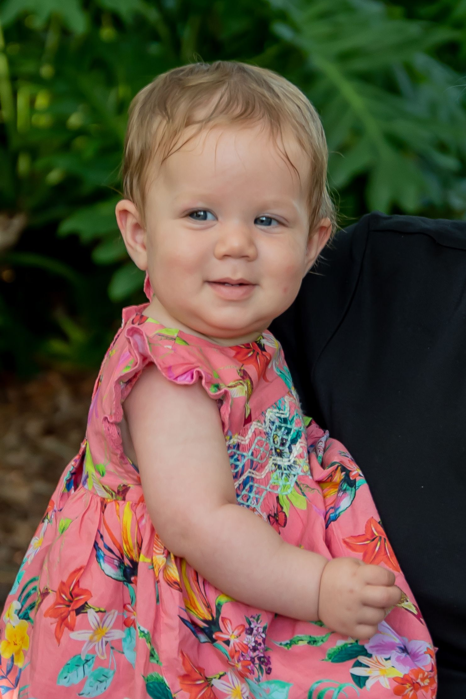 Smiling baby girl in pink floral dress standing outdoors with green tropical plants in background.