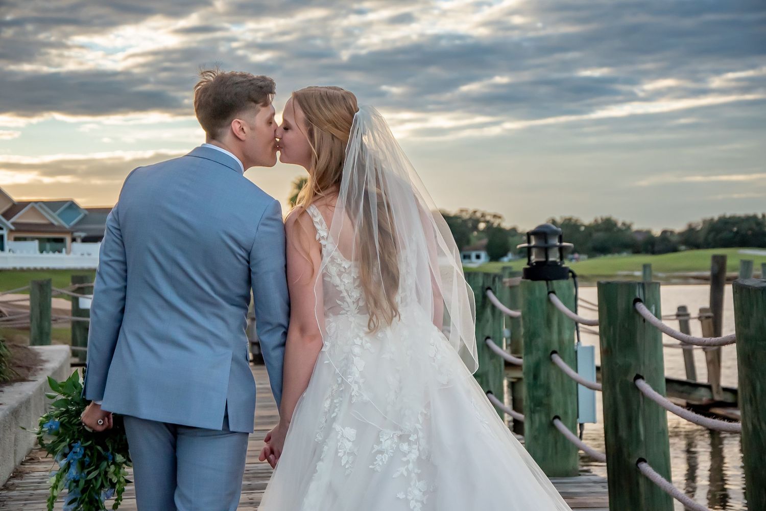 Bride and groom share a romantic kiss on a waterfront dock at sunset, bride wearing lace gown and veil.