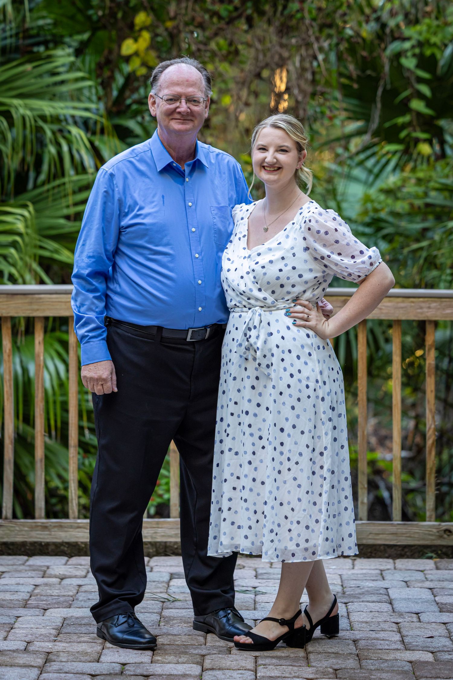 A man in a blue shirt and woman in a white polka dot dress pose together outdoors on a brick patio.