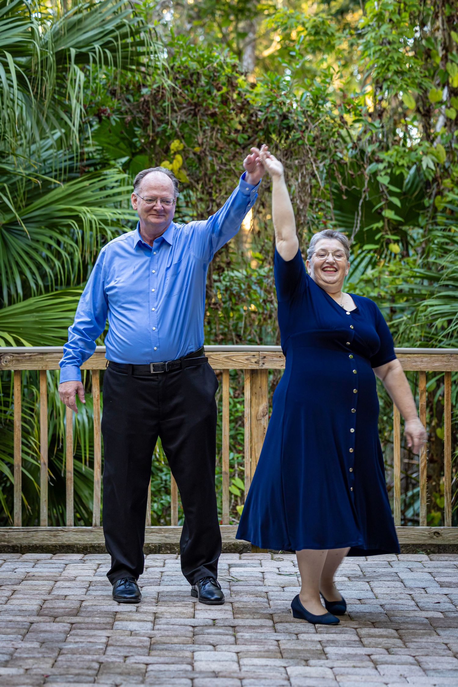 Senior couple dancing outdoors on brick patio, holding hands raised, surrounded by lush tropical greenery.