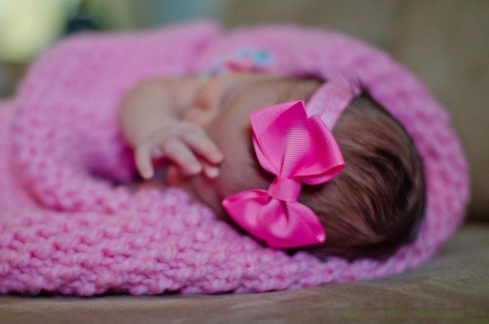 Newborn baby girl sleeping wrapped in a pink knit blanket with a pink bow headband.