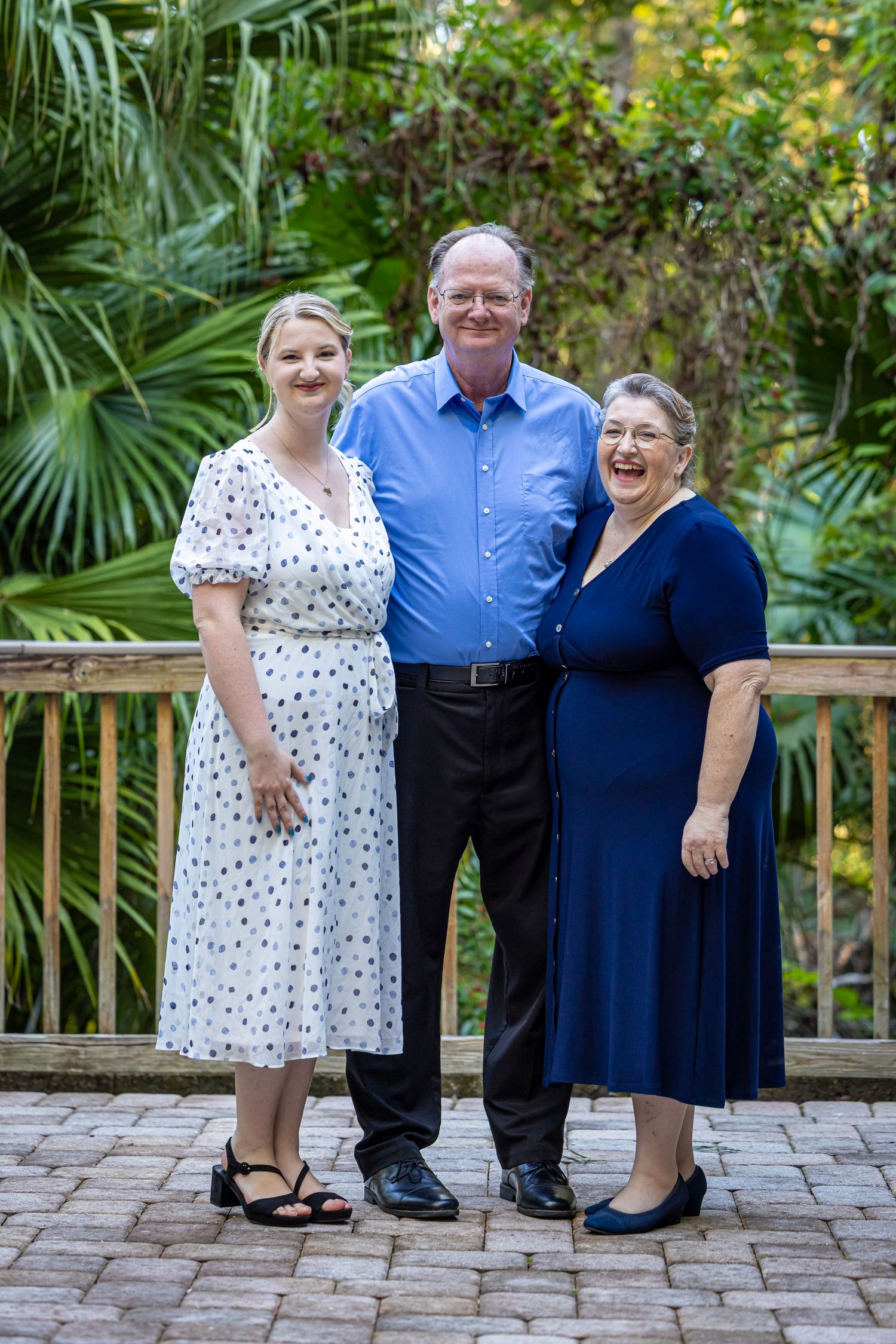 Three smiling people pose together on a brick patio surrounded by lush tropical greenery.