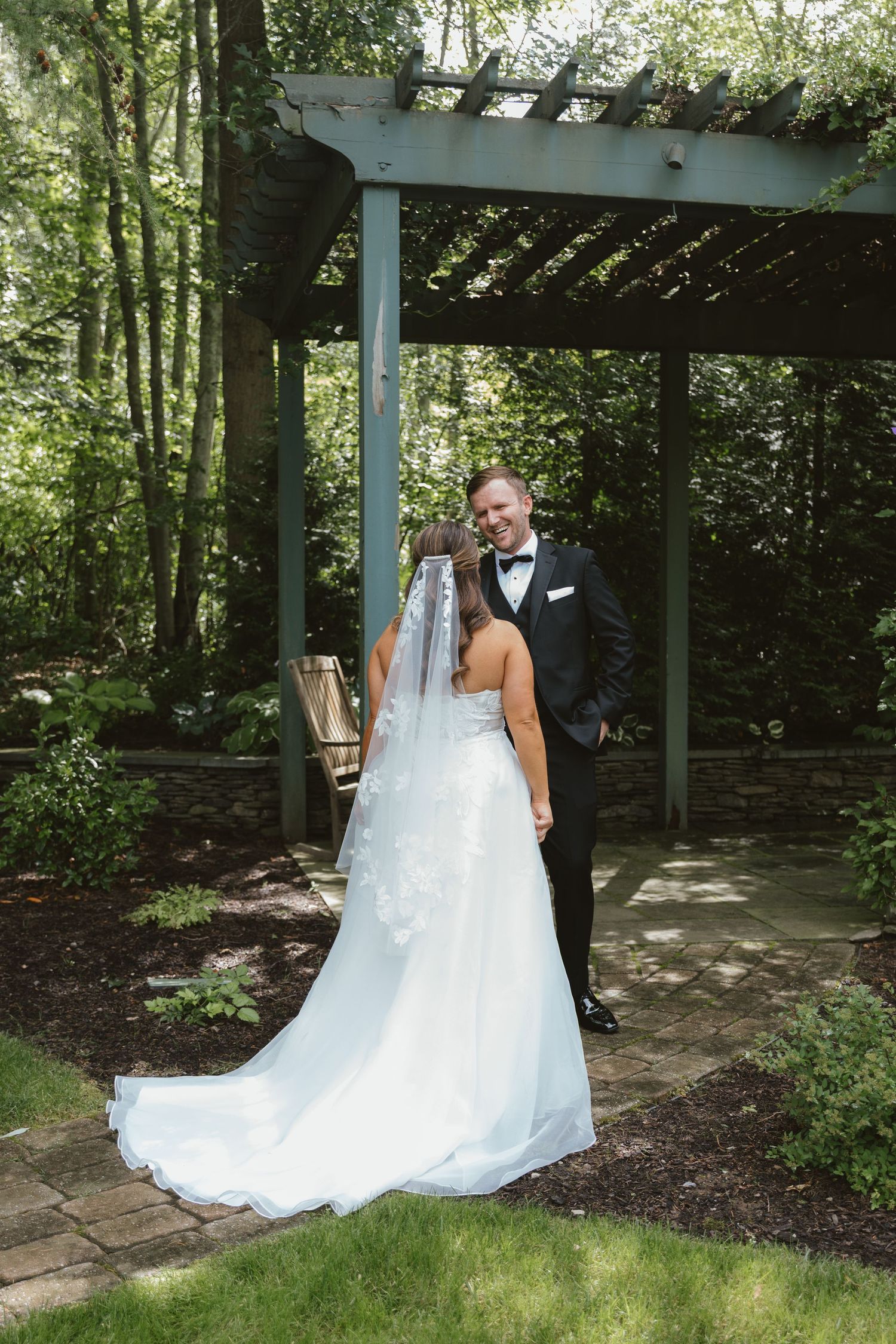 Bride in white gown with veil and groom in black tuxedo posing under a wooden pergola in a lush garden setting.