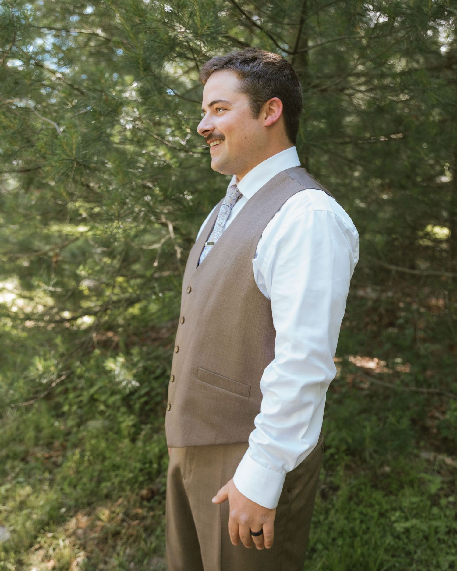 Smiling groom in brown vest and white dress shirt standing outdoors among green trees.