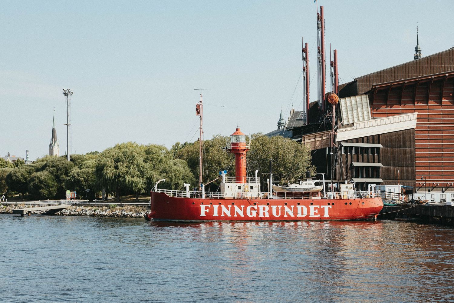The red lightship Finngrundet moored along a sunny Stockholm waterfront with historic buildings behind.