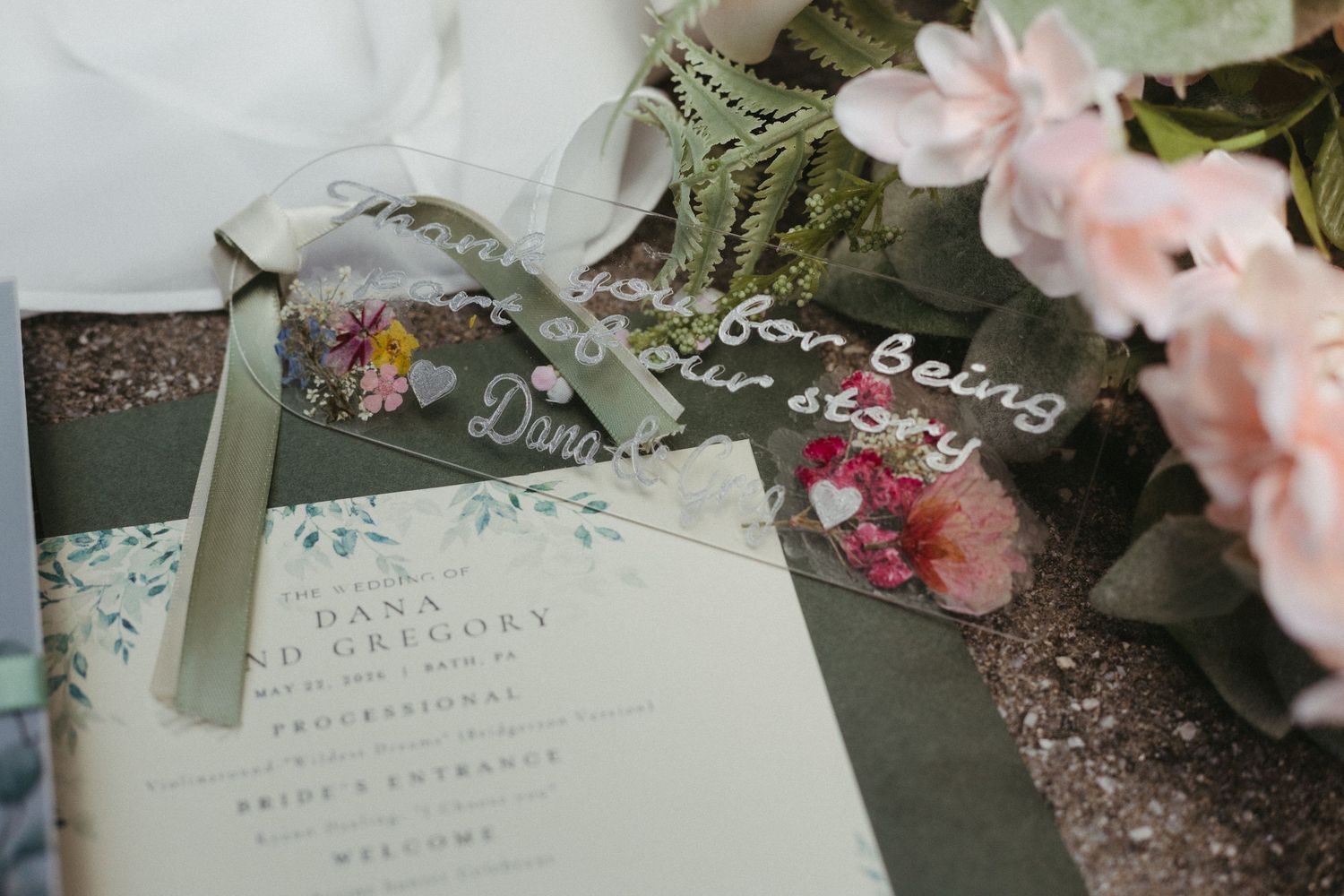 Elegant wedding stationery flat lay featuring floral programs, acrylic signage, and sage green ribbons with pink blooms.