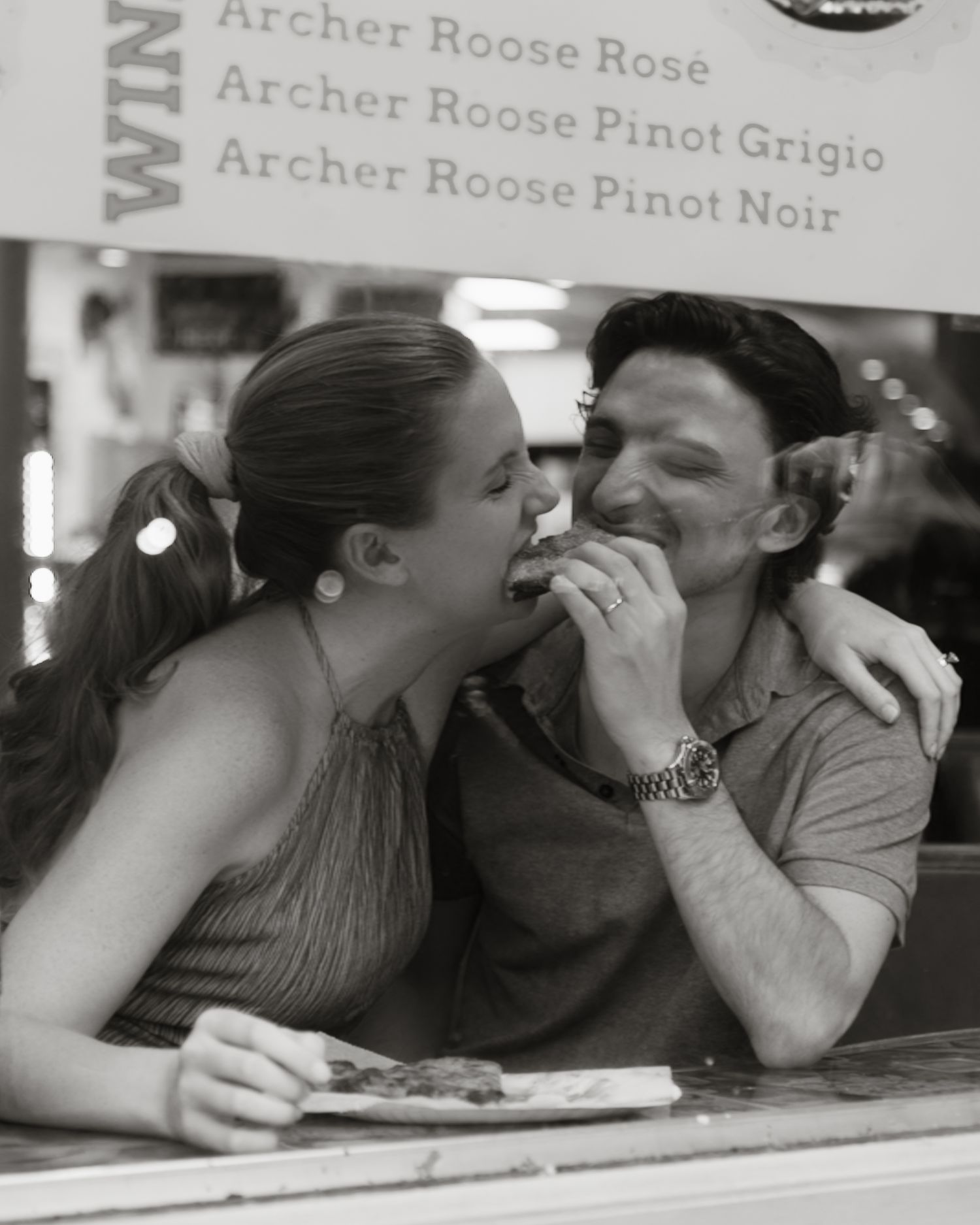 Black and white photo of couple playfully sharing food at a wine bar window.
