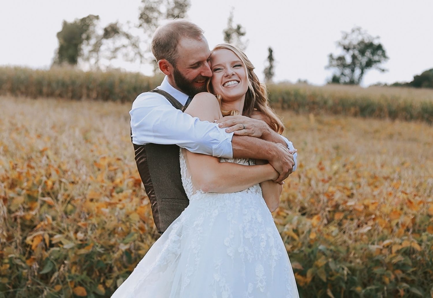 Groom hugs smiling bride in tulle gown in golden autumn field surrounded by fall foliage.