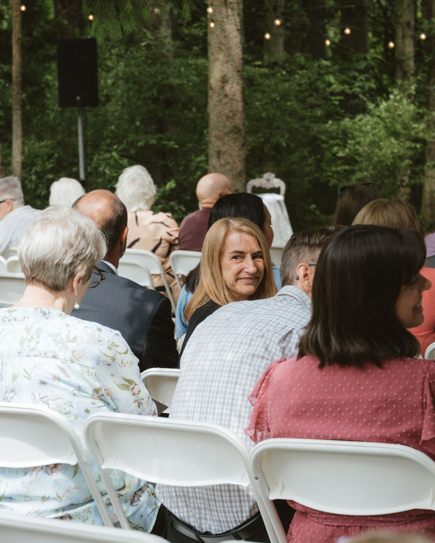 Wedding guests seated in white chairs outdoors under string lights in wooded ceremony setting.