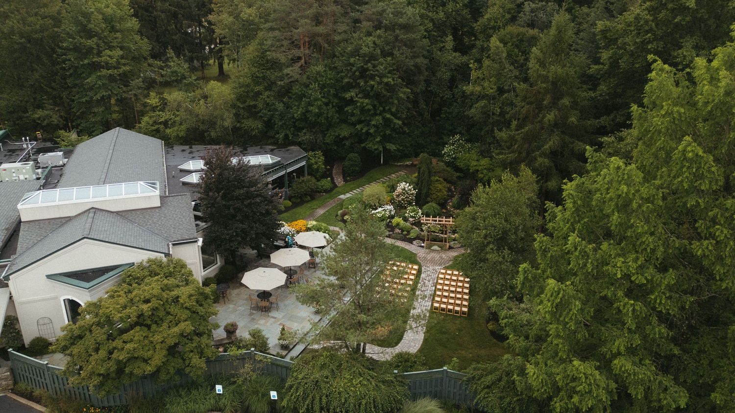 Aerial view of a lush garden venue with outdoor seating, umbrella tables, and surrounding trees.