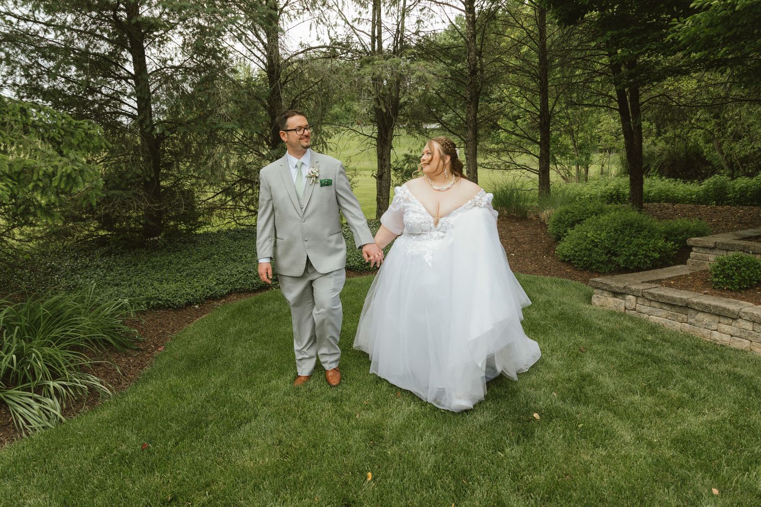 Bride and groom walk hand in hand through lush green garden path on their wedding day.