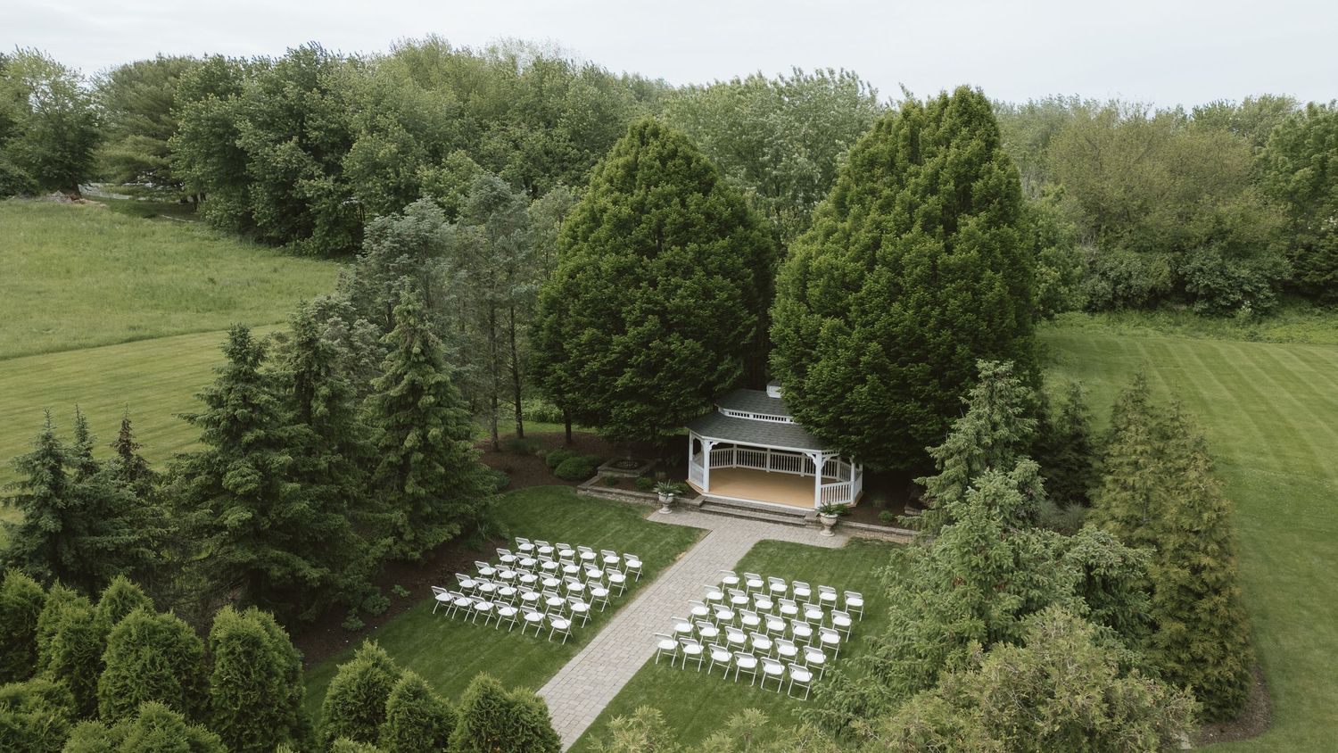 Aerial view of an outdoor wedding ceremony setup with white chairs arranged near a gazebo surrounded by lush green trees.